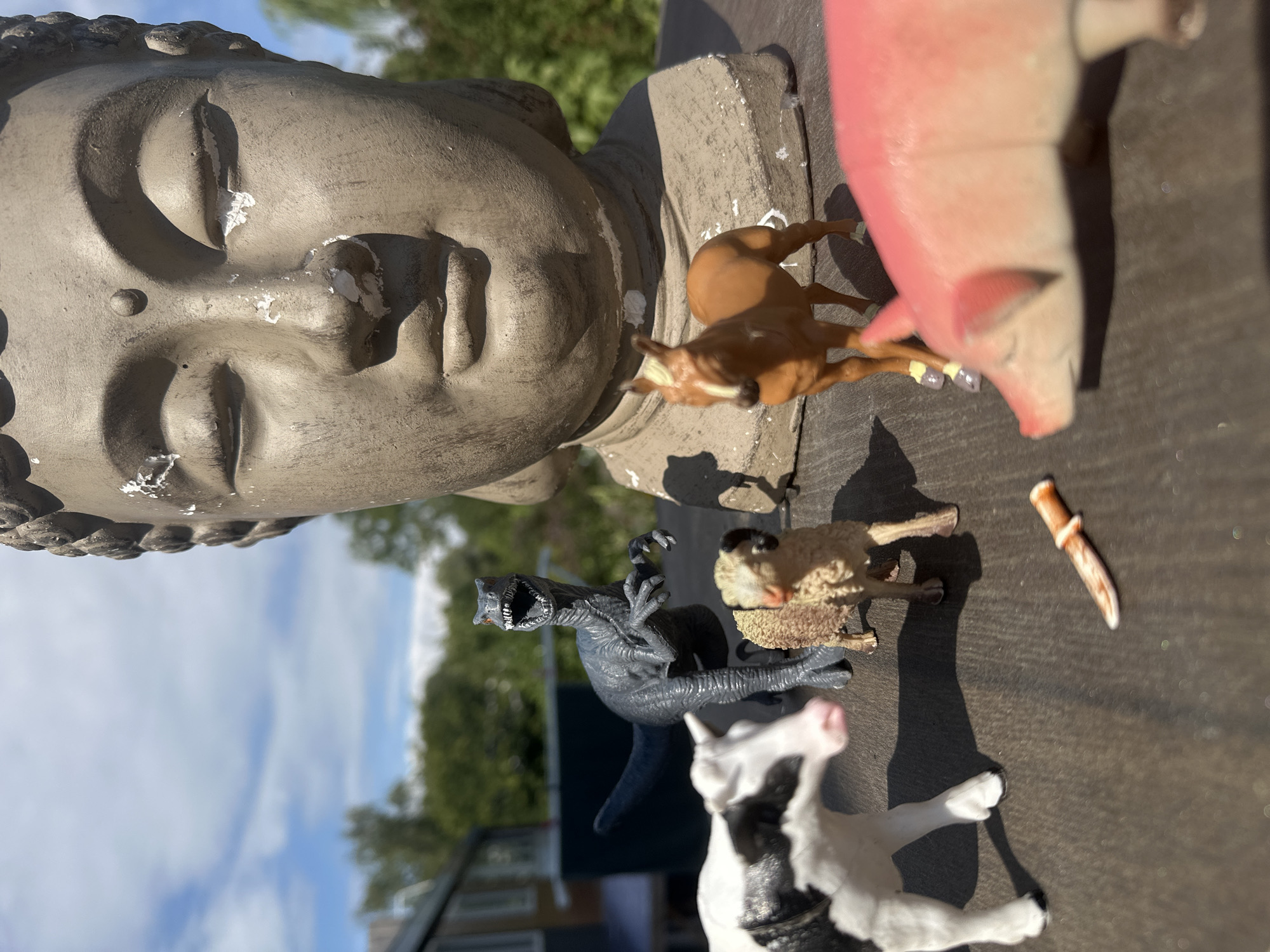 The image depicts a collection of toy animals and a statue of a Buddha's head arranged on a weathered wooden surface, likely a table or deck, outdoors. A dark grey dinosaur toy stands alert, its mouth open, facing a black and white cow toy. To the right of the cow is a small, tan sheep toy with black horns. Further to the right, a brown horse toy stands near the base of the Buddha statue, which is weathered and shows signs of paint chipping. In the foreground, a pink pig toy is partially visible, and a small, orange object resembling a miniature knife lies on the surface between the pig and the sheep. The background consists of green foliage, trees, and a residential building under a partly cloudy blue sky. The lighting suggests it is daytime. This scene is not in Lelystad, Netherlands.
