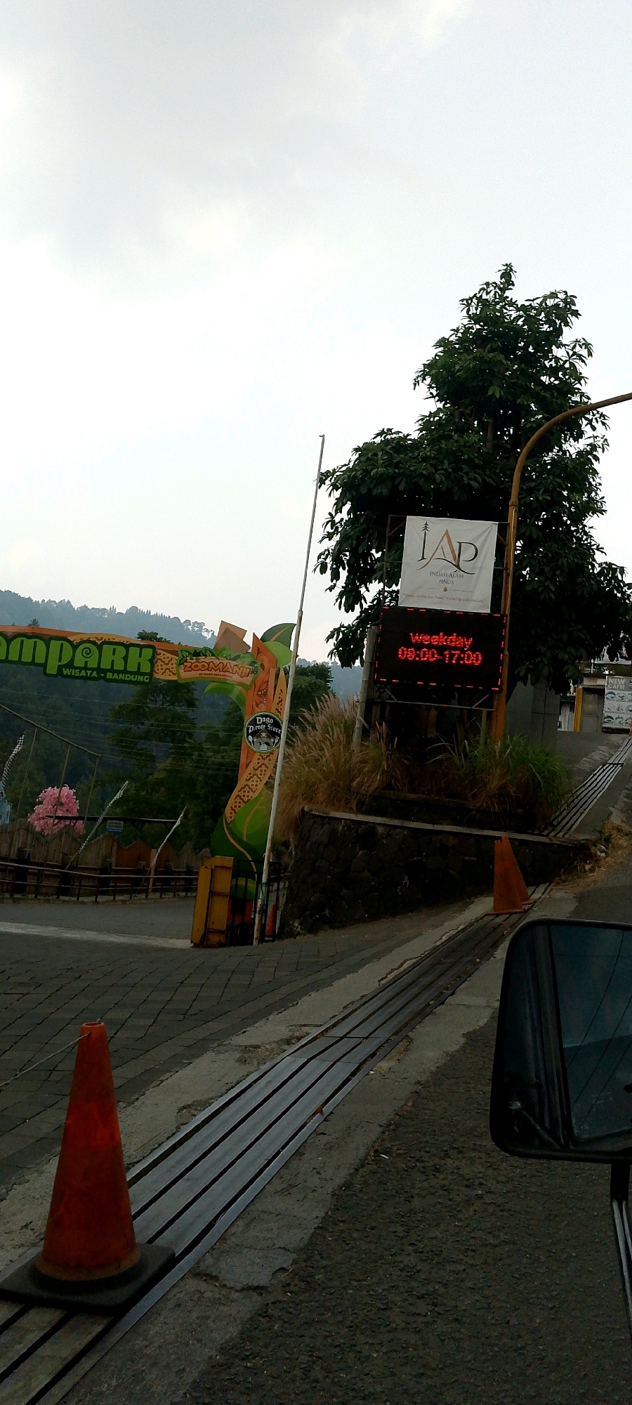 The image captures an outdoor scene, likely near an entrance to an attraction or park. A large, colorful archway with "ZOOMANJI" and "WISATA - BANDUNG" written on it is visible on the left. A traffic cone, orange in color, stands on the paved road in the foreground. To the right, a large tree with dark green foliage is prominent. Behind the tree, a digital sign displays "weekday" and "09:00-17:00" in red LED lights. Another banner with the logo "IAP" and "INDAM ALAM PINUS" is also present. Beyond the immediate entrance area, a hilly, green landscape with some buildings can be seen. The sky is overcast. The overall impression is that of an entrance to a recreational area on a cloudy day. The specific location of Mekarwangi, Indonesia is not visually identifiable from the image itself.
