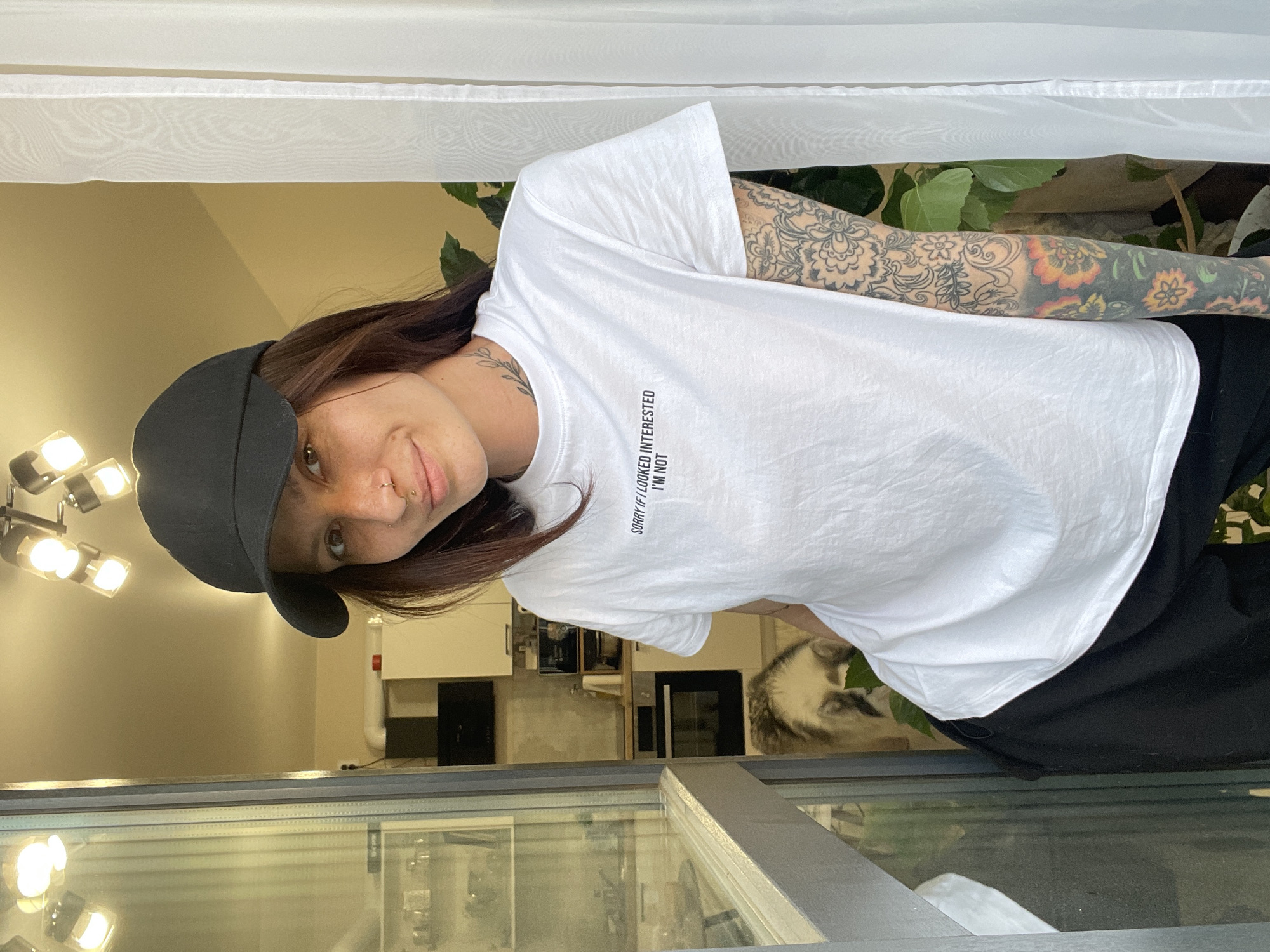 The image depicts a person wearing a black baseball cap and a white t-shirt with the text "SORRY IF I LOOKED INTERESTED I'M NOT". The person has brown hair, fair skin, and facial piercings. The person has tattoos on their left arm. The location appears to be indoors, possibly a residence with a kitchen visible in the background with kitchen cabinets and an oven. A dog is also present in the background. The location is purportedly in the city of Pervomaysky, Russia, though this cannot be visually confirmed within the image. The person is near a glass-covered structure.