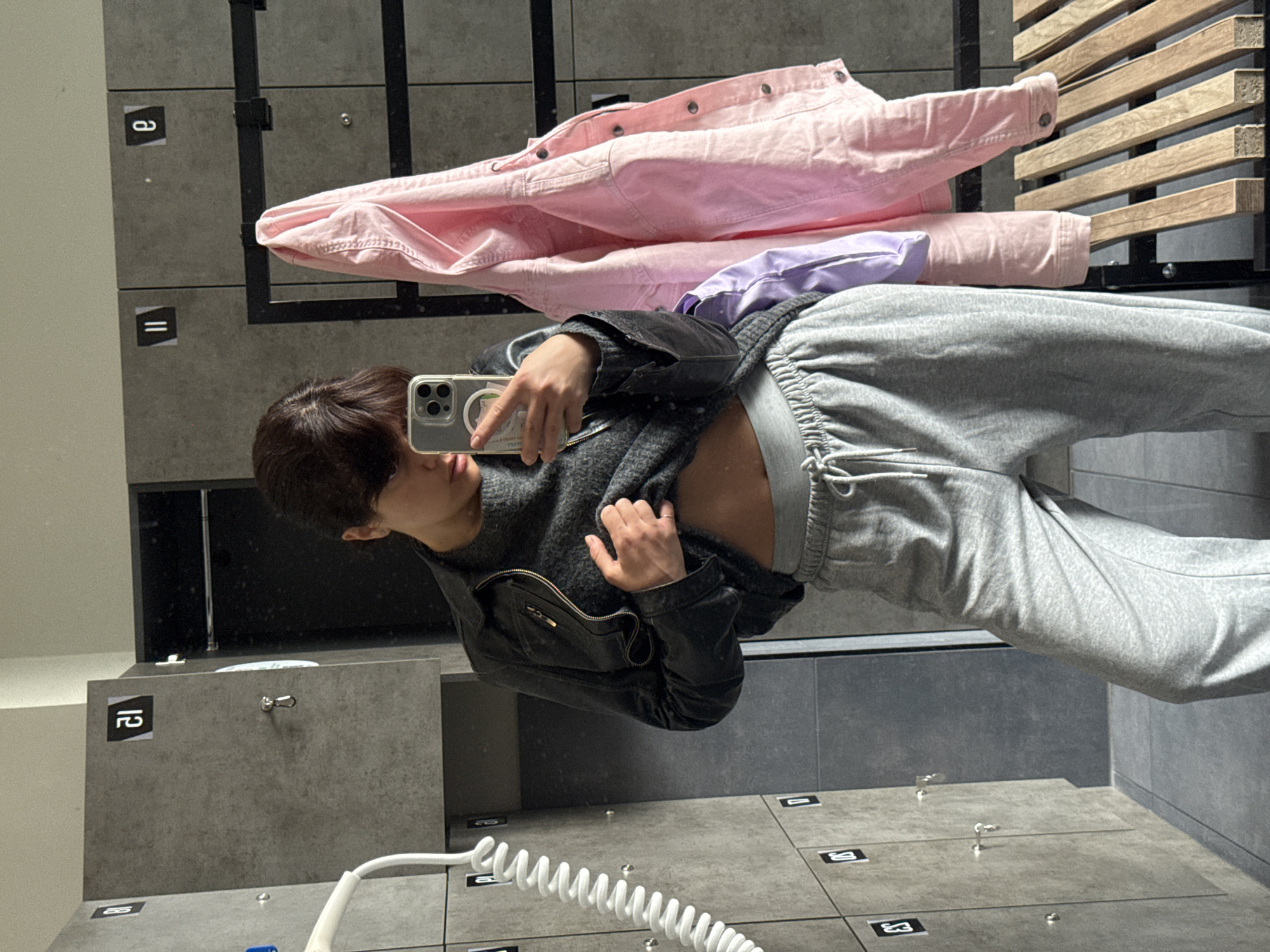 The image shows a person taking a selfie in front of a bank of lockers. The person is wearing gray sweatpants, a dark sweater, and a black jacket. A pink jacket and purple shirt are visible on top of the lockers. The lockers appear to be in a public facility, possibly a gym or a locker room. There are numbers on the lockers and a white curly cord hanging down. The location cannot be determined from the image with certainty, but given the style of the lockers and the general aesthetic, it's plausible to be in Berlin, Germany or somewhere else in Europe.