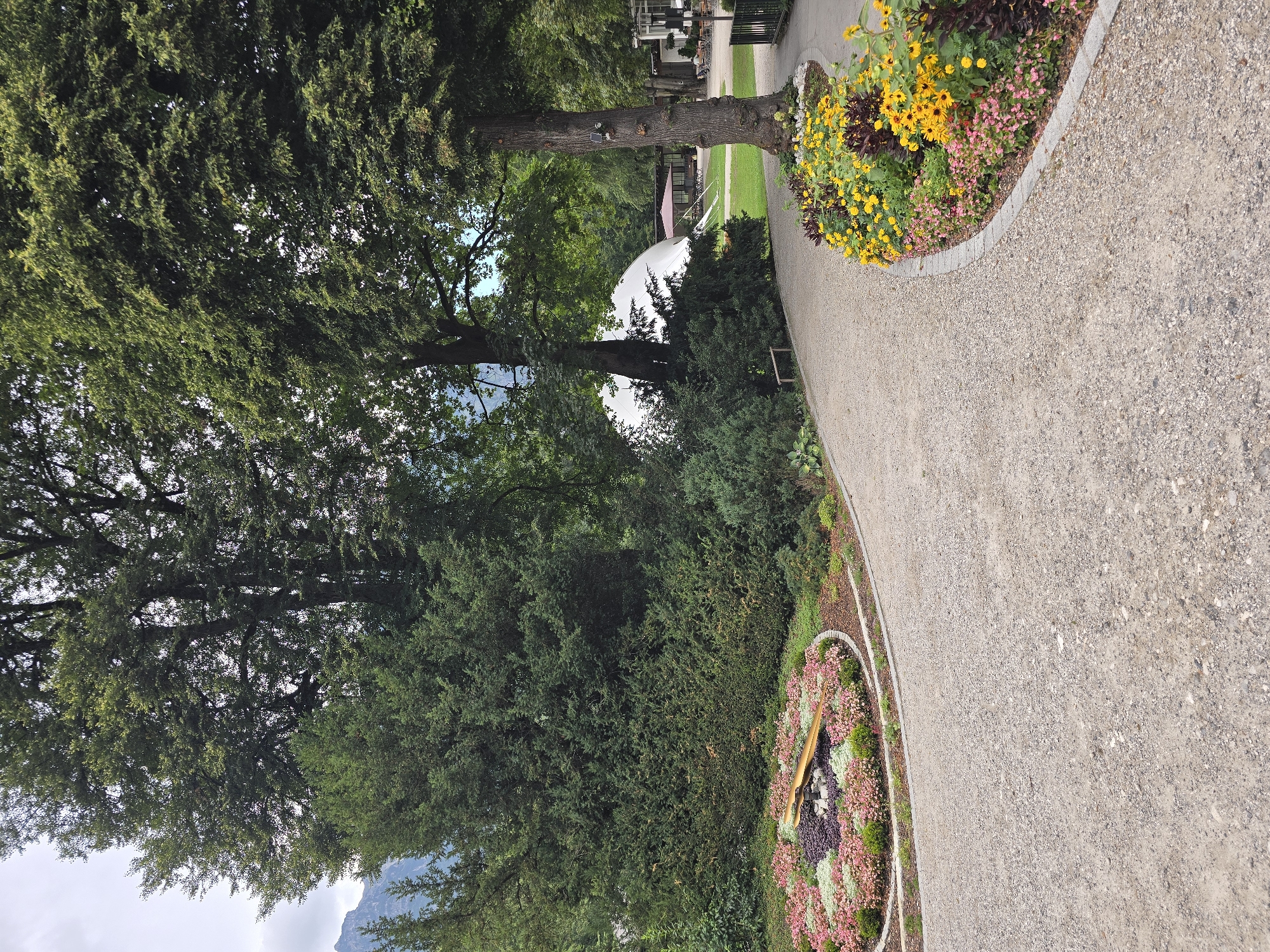 The image shows a park pathway in Garmisch-Partenkirchen, Germany. The pathway is made of gravel and curves to the right. On the left side of the pathway, there is a circular flower bed designed to look like a clock face with golden hands. Behind the flower bed are lush green trees and shrubs. On the right side of the pathway, there is another flower bed with yellow and pink flowers. In the background, a large white dome structure is visible, along with some outdoor seating and umbrellas, suggesting a cafe or event space. The environment appears to be a well-maintained public park. There are no people clearly visible in the image.