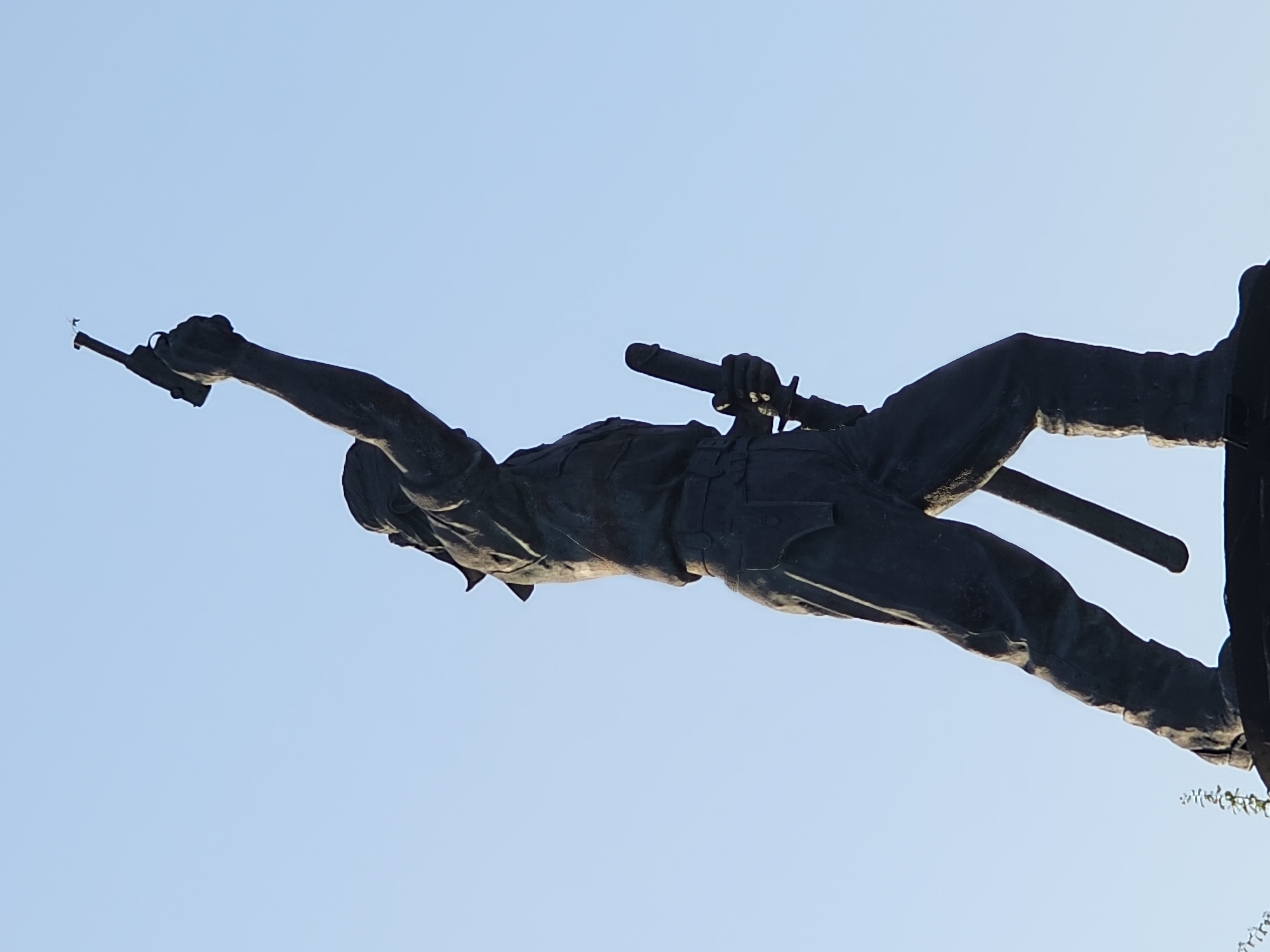 This is a low-angle, medium shot of a bronze statue of a man holding a pistol in his raised right hand and a sword in his left hand. The statue is dark in color and appears weathered. The man is dressed in what looks like military attire, with a bandana or headscarf tied around his head, a buttoned shirt with pockets, and trousers tucked into boots. He is standing on a dark base, possibly a pedestal, which is only partially visible. The background is a clear, pale blue sky, suggesting it is daytime. The statue is likely located in the city of Denpasar, Indonesia, as it is a memorial to I Gusti Ngurah Rai, a Balinese hero who led resistance against the Dutch. The statue is a prominent landmark in Denpasar and is often referred to as the "Patung Puputan" or "Patung I Gusti Ngurah Rai".
