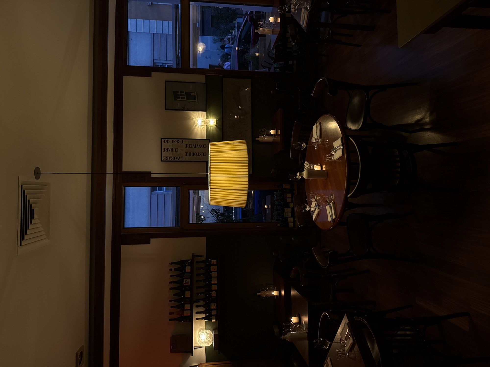 The image depicts the interior of a restaurant with a warm, dimly lit ambiance. Wooden tables are set with glassware and cutlery. A large, yellow lampshade hangs from the ceiling, casting a soft glow. Shelves lined with wine bottles are visible against a dark wall. In the background, an open doorway reveals a glimpse of the street outside, where a red car is parked, and several people are standing. The restaurant appears to be located in Belgrade, Serbia, given the presence of Cyrillic text on some of the posters visible on the wall. The scene suggests a relaxed evening setting, possibly after business hours or during a quiet period.