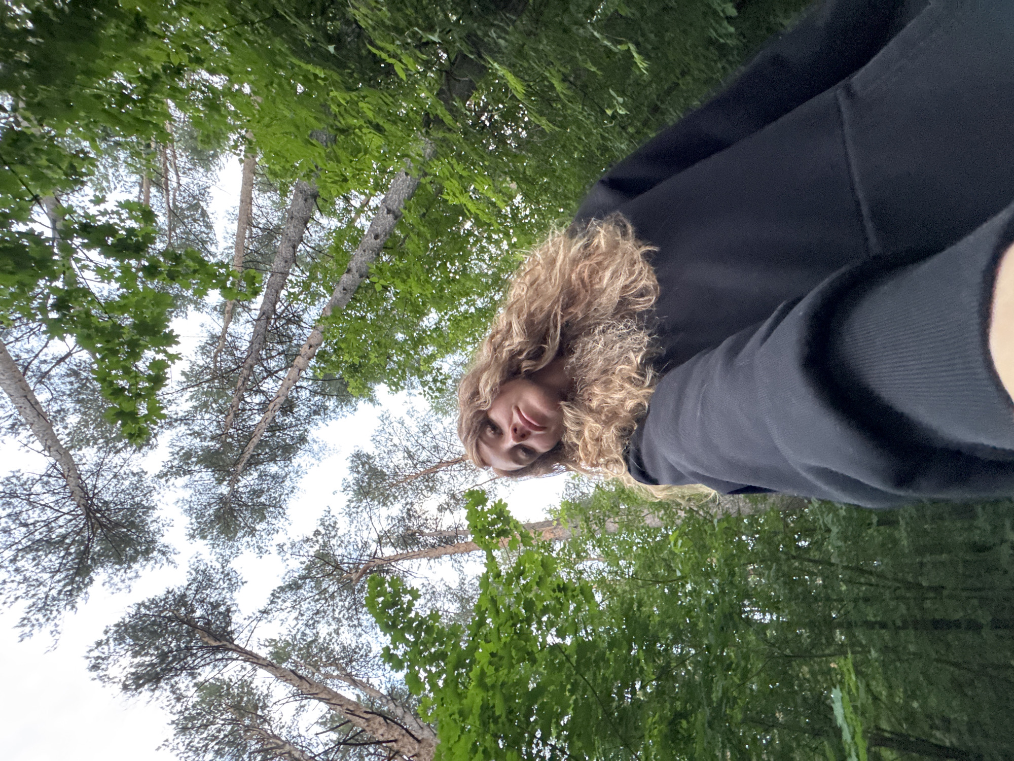 A low-angle, wide shot captures a young woman with long, curly blonde hair in a black hoodie, holding a phone to take a selfie. She is positioned in the lower right of the frame, looking up and slightly towards the camera. The background is a dense forest with tall, slender trees whose canopies reach towards a bright, overcast sky. Lush green foliage fills the spaces between the trees. The image appears to have been taken in a natural, wooded environment.
