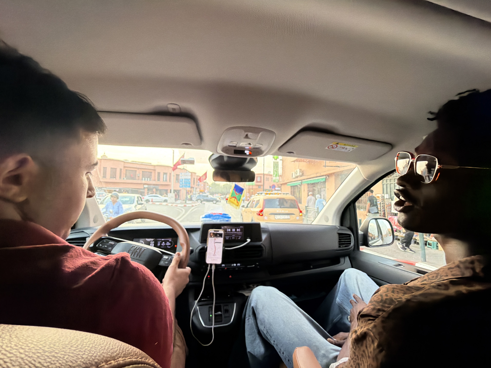 Two people are inside a vehicle, likely a passenger van, in Marrakesh, Morocco. The person on the left, wearing a maroon shirt, is driving and holding the steering wheel. The person on the right, wearing glasses and a patterned shirt, is in the passenger seat, looking to the right. The dashboard features a navigation screen on a smartphone and a car's infotainment system. Outside the vehicle, a street scene unfolds with buildings, other vehicles, and pedestrians, characteristic of a Moroccan city. The architecture suggests a historic urban environment. The lighting indicates it is daytime.
