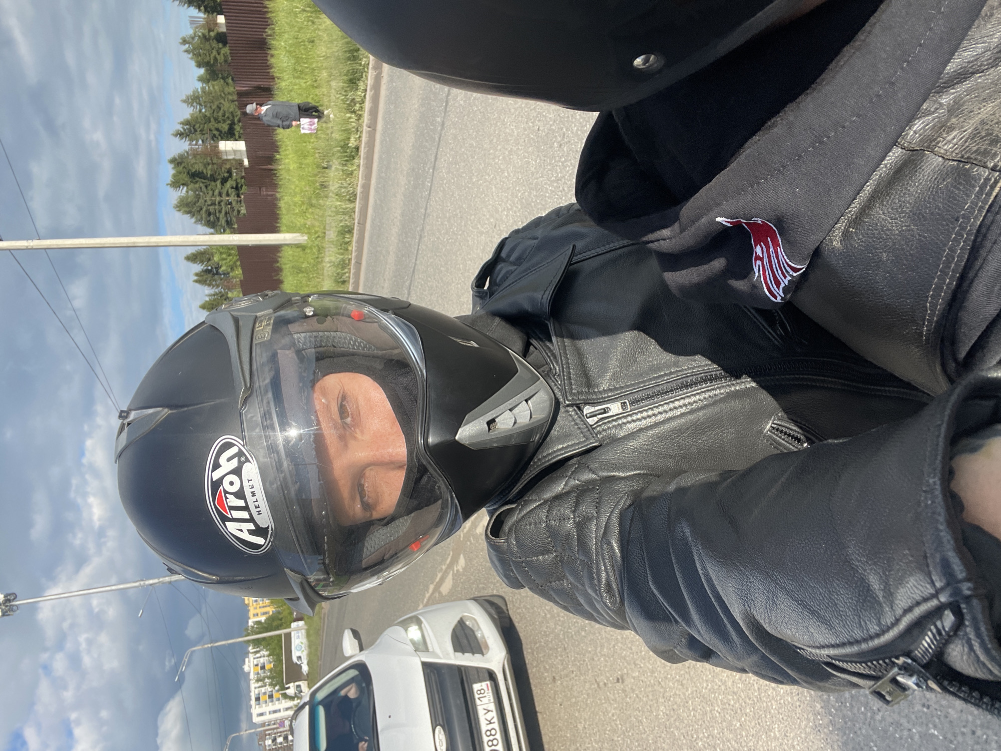 The image, correctly oriented, shows a first-person perspective, likely a selfie, of a person on a motorcycle or as a passenger. The main subject is wearing a black Airoh brand motorcycle helmet, with their eyes and upper face visible through the clear visor. They are also wearing a black leather jacket with quilted details on the shoulders and arms, and prominent zippers. Underneath the jacket, a black garment with a distinctive red and white emblem, resembling a stylized bird or abstract design, is visible.
The scene depicts an outdoor setting on a paved road during the daytime. The sky is partially cloudy but bright, indicating sunny weather. To the left side of the road, there's a grassy verge, a brown wooden fence, and several evergreen trees. A lone pedestrian, dressed in a hat and jacket and carrying a bag, is visible walking near the fence in the distance. To the right, further down the road, white buildings, possibly residential, are visible against the skyline.
In the foreground and midground, another white car is partially visible ahead of the motorcycle, bearing a Russian license plate "X88KY 18 RUS". The "18 RUS" code indicates the Udmurt Republic, aligning with the provided location context of Izhevsk, Russia. A utility pole stands on the left side of the road. The overall impression is a casual ride through a semi-urban or suburban area. The person's expression behind the visor appears neutral.