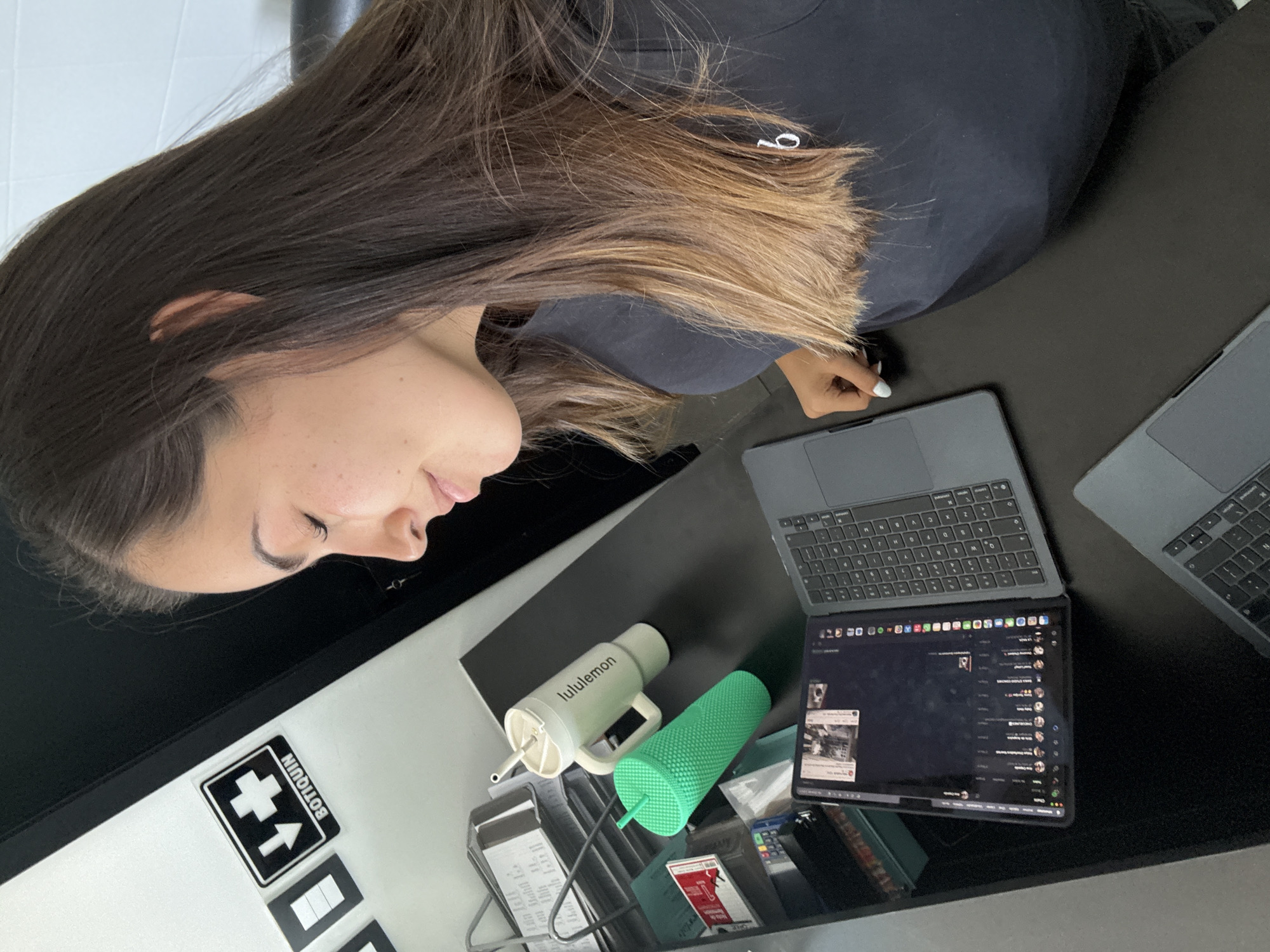 An indoor scene captures a young woman seated at a dark desk, engaged with electronic devices. She is seen from the side, looking down, with long, dark brown hair that transitions to lighter, golden-brown highlights towards the ends. She wears a dark-colored top, possibly a sweatshirt, with a small white embroidered logo visible on the chest. Her hand, adorned with light-colored nail polish, rests near one of the laptops.
On the dark desk, two laptops are present. The one in the foreground is open, displaying a screen that appears to be a messaging application, showing a list of contacts and chat content. To its right, a second laptop is partially visible. Also on the desk are two distinct drinking containers: a light green "Lululemon" branded bottle with a white lid, handle, and straw, and a vibrant green studded tumbler with a straw. A clear desk organizer holds various papers or receipts and a red card.
In the background, to the left of the woman, a white surface with what appears to be white tiles is visible. Affixed to another white surface, possibly a wall or cabinet, is a black and white sticker featuring a first aid cross symbol, an upward arrow, and the Spanish word "BOTIQUIN" (First Aid Kit) written vertically on the right side. The overall lighting suggests it is daytime. The woman appears focused, possibly working or browsing. The presence of the "BOTIQUIN" sign aligns with the provided location context of San Bernardino Tlaxcalancingo, Mexico.