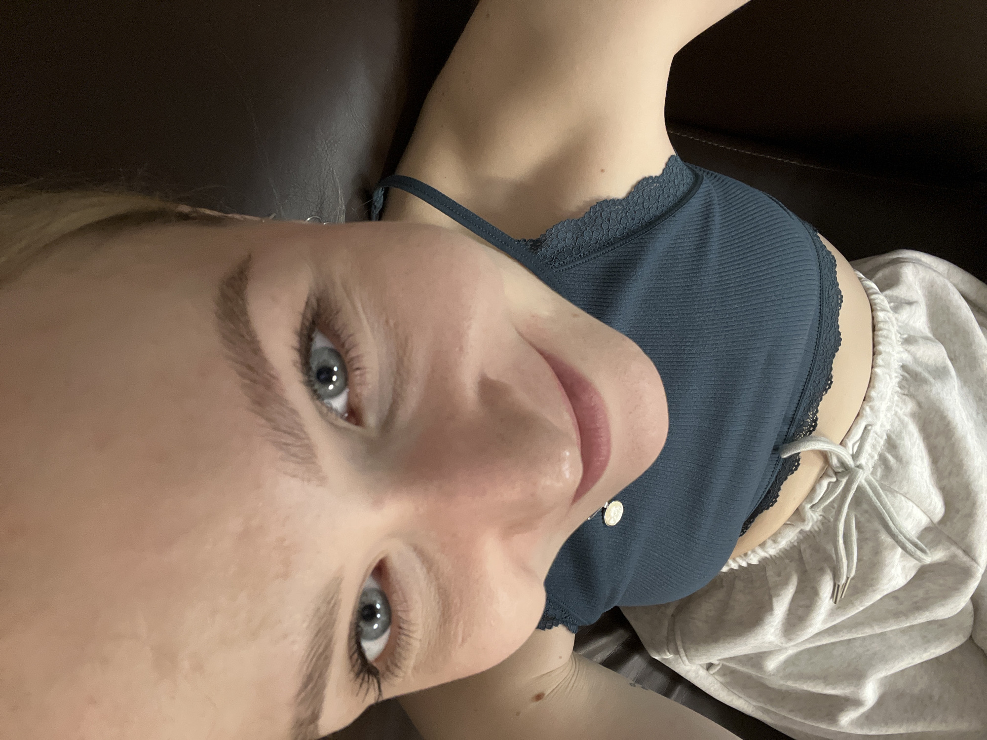 A close-up, selfie-style image shows a woman with fair skin, light eyebrows, and blue eyes. She is wearing a teal-colored tank top with lace trim and light grey sweatpants with a drawstring. The background appears to be a dark brown leather surface, possibly a couch. The woman is angled towards the camera, with her face prominently featured. There is no indication of a location in Khimki, Russia.