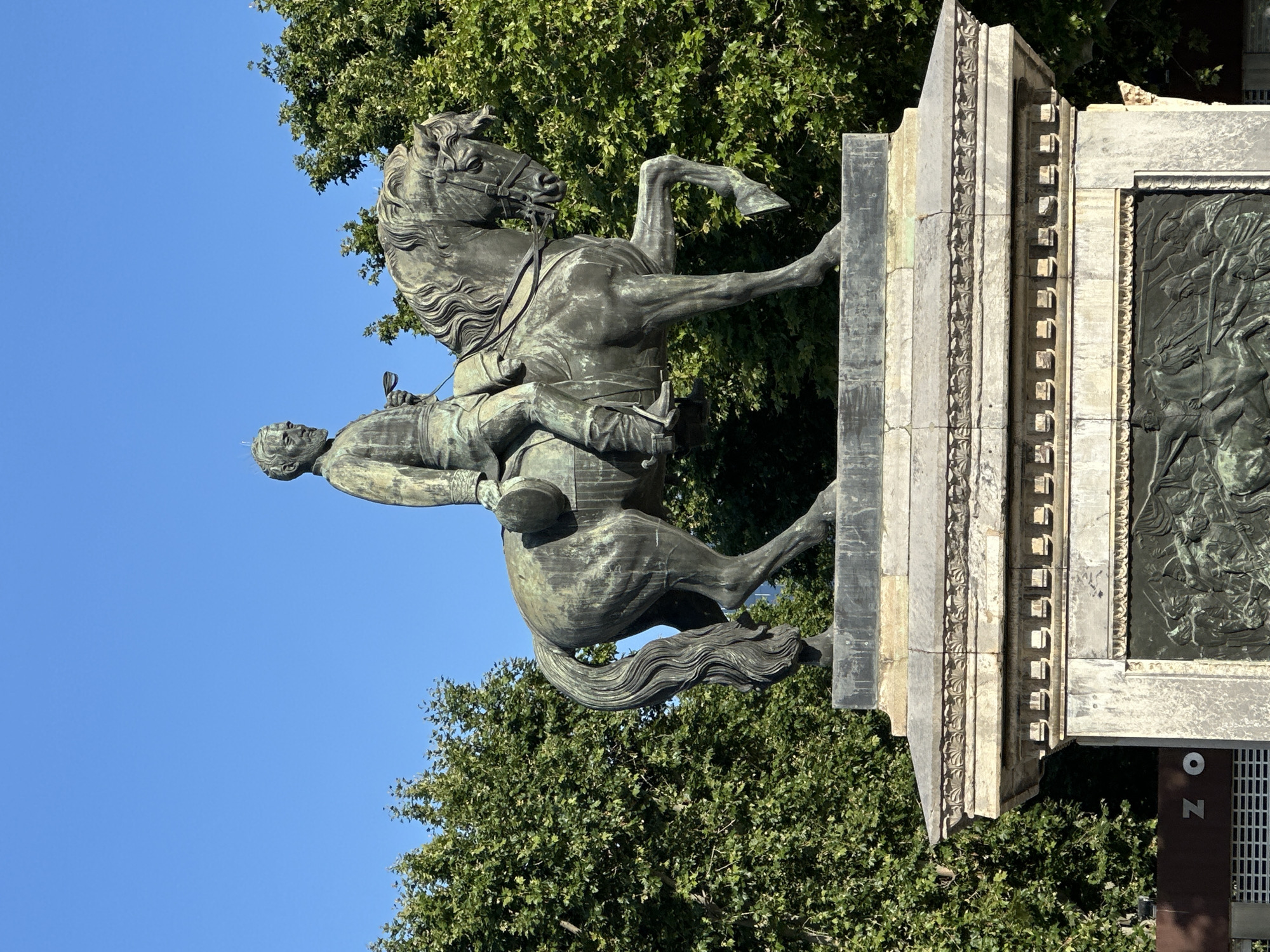 A bronze equestrian statue of General Joan Prim i Prats stands prominently against a clear blue sky, surrounded by vibrant green trees. The statue depicts General Prim, a bearded man in a detailed military uniform, seated on a horse with his right hand holding a small object, possibly a whip or baton. The horse is captured in a dynamic pose, with its left front leg raised. The statue rests on an ornate, multi-tiered stone pedestal featuring a cornice with dentil molding and, on its visible side, a bronze bas-relief depicting a battle scene with multiple figures on horseback and on foot engaged in combat. The scene is illuminated by bright daylight, casting subtle shadows on the statue's details. In the bottom left corner, part of a modern building with the letters "Z O" visible suggests proximity to the Barcelona Zoo. The entire scene is located within Parc de la Ciutadella in Barcelona, Spain.