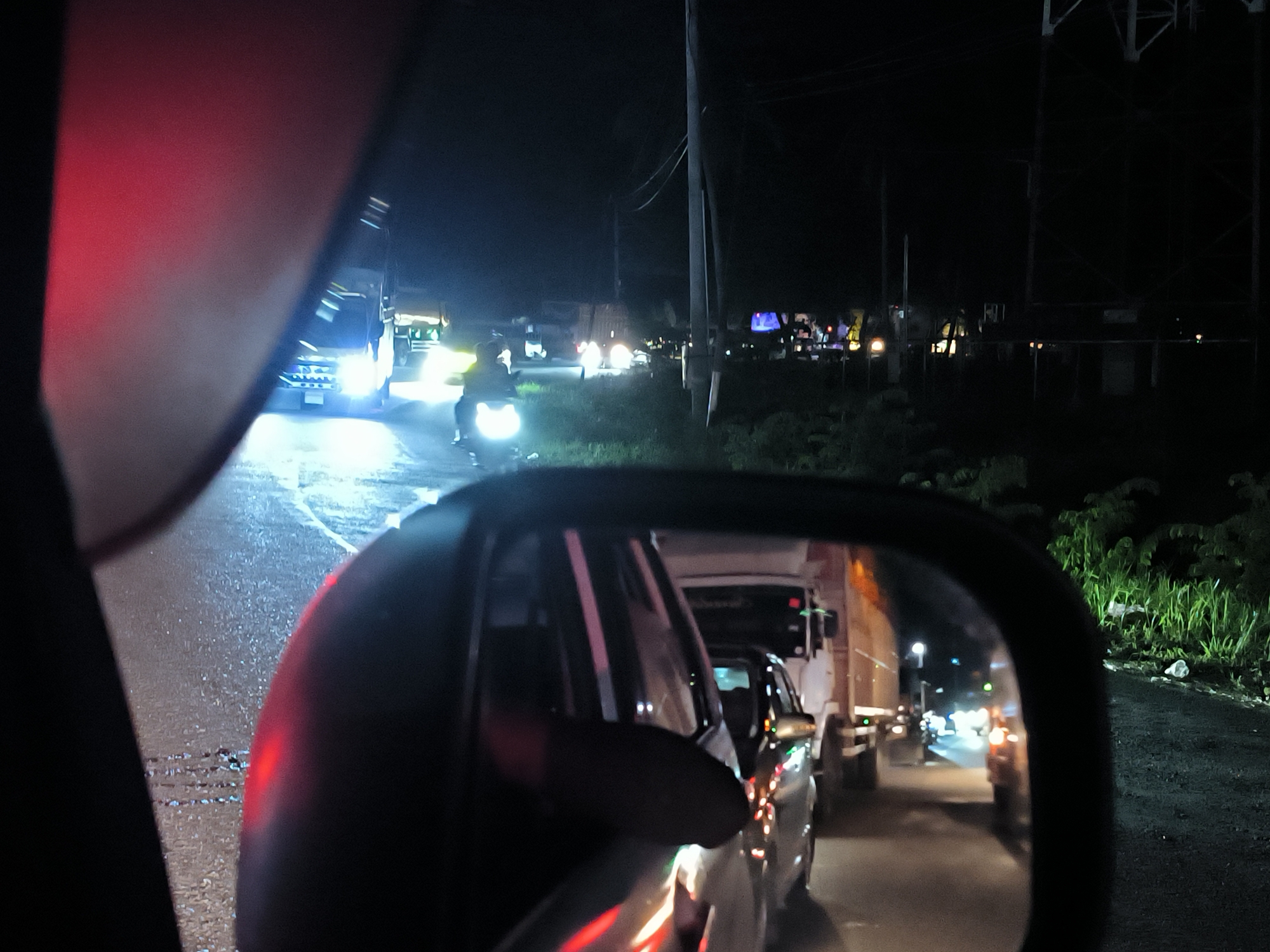 The image shows a night scene in Keudetanjong, Indonesia, likely taken from inside a vehicle. The focus is on the road ahead and the view reflected in the car's side mirror.
In the distance, there's a motorcycle with a bright headlight and a white van. Cars and trucks are lined up behind the van and in the side mirror. There are power lines on the right side of the image, along with roadside vegetation and litter. The general environment suggests a busy road or traffic congestion.