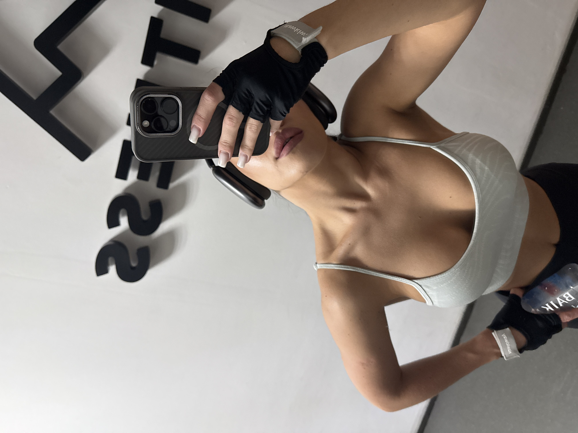 A young woman in workout attire is taking a selfie in front of a white wall with large black lettering. She wears a light-colored sports bra, black workout gloves, and black leggings. She holds a phone up to take the picture, obscuring the lower portion of her face. A bottle of water is visible in her other hand. The image appears to be taken in a fitness center, based on the attire and the presence of the water bottle. The lettering on the wall could be part of the fitness center's branding. The water bottle has the word "Baikal" written on it. The overall setting appears to be modern and clean, consistent with the style of a contemporary gym. Due to the text on the water bottle, it is implied that the image may be taken in Russia. No specific information or elements in the picture indicate that this image was taken in Kazan.