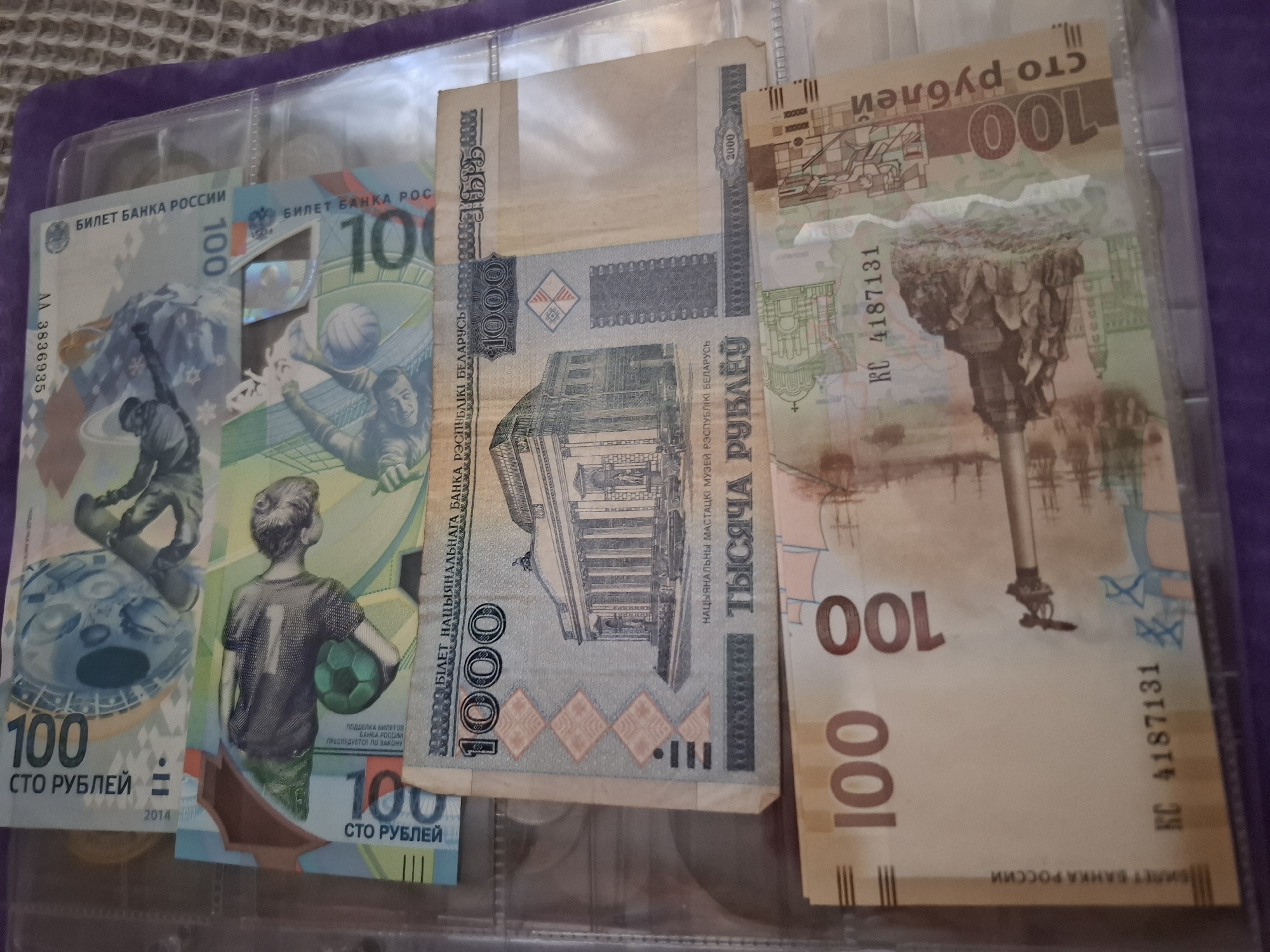 The image shows a collection of banknotes from Russia and Belarus displayed in a plastic binder. There are two 100 Russian Ruble banknotes, one showing a child in a football shirt, and another one depicting a man on a snowboard, both issued in 2014. There's also a 1000 Belarusian Ruble banknote, and a 100 Russian Ruble banknote with a vertical orientation, showing a monument. The image does not suggest any connection to the city of Amzya, Russia.