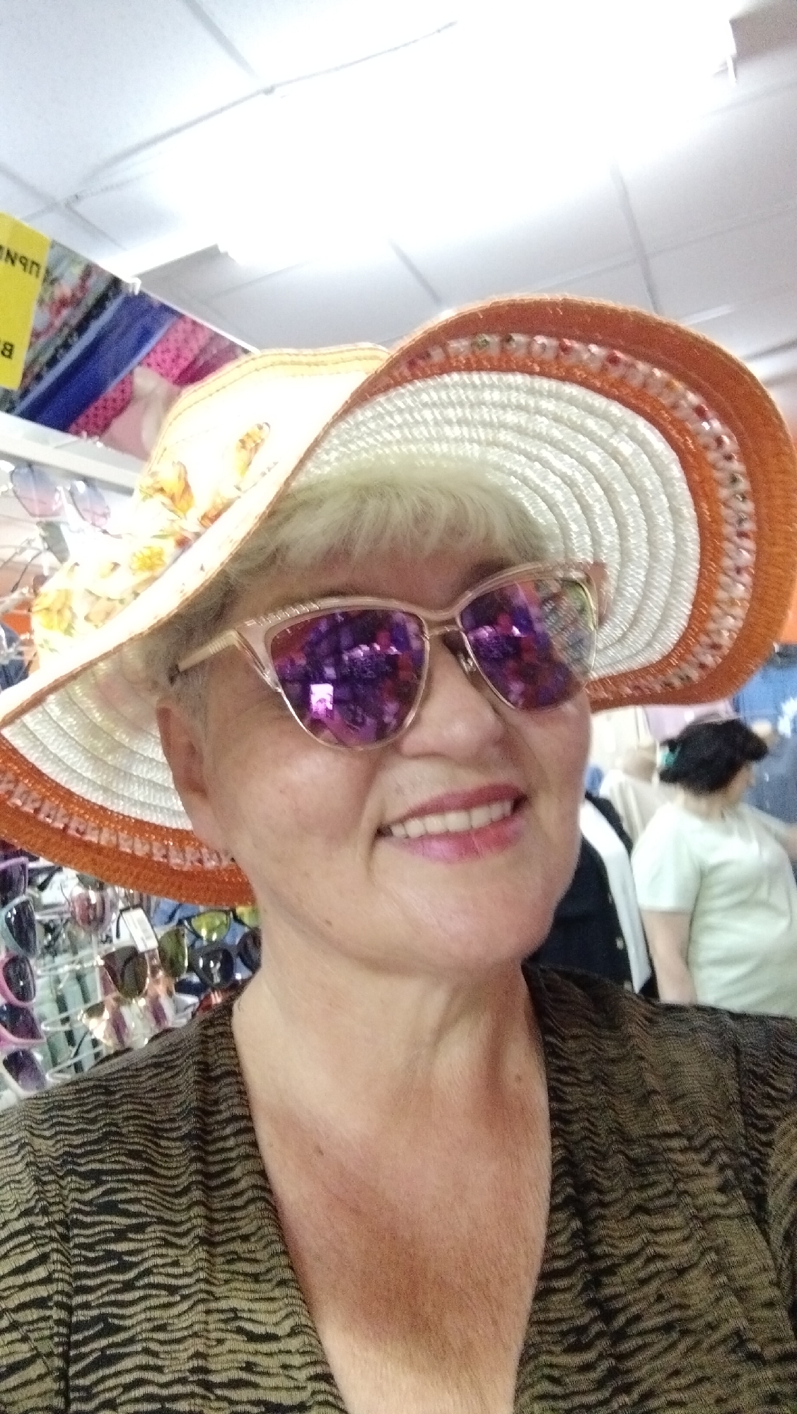 An older woman with short, blonde hair is shown in a close-up, selfie-style shot. She is wearing a wide-brimmed hat with orange trim and a floral embellishment. Her sunglasses have pink frames and purple lenses that reflect the surroundings. She is also wearing a dark, patterned top. In the background, several racks of sunglasses are visible, suggesting the photo was taken in a store. Another woman with dark hair is partially visible in the background to the right. The image appears to have been taken indoors, possibly in a shop in Horishni Plavni, Ukraine.