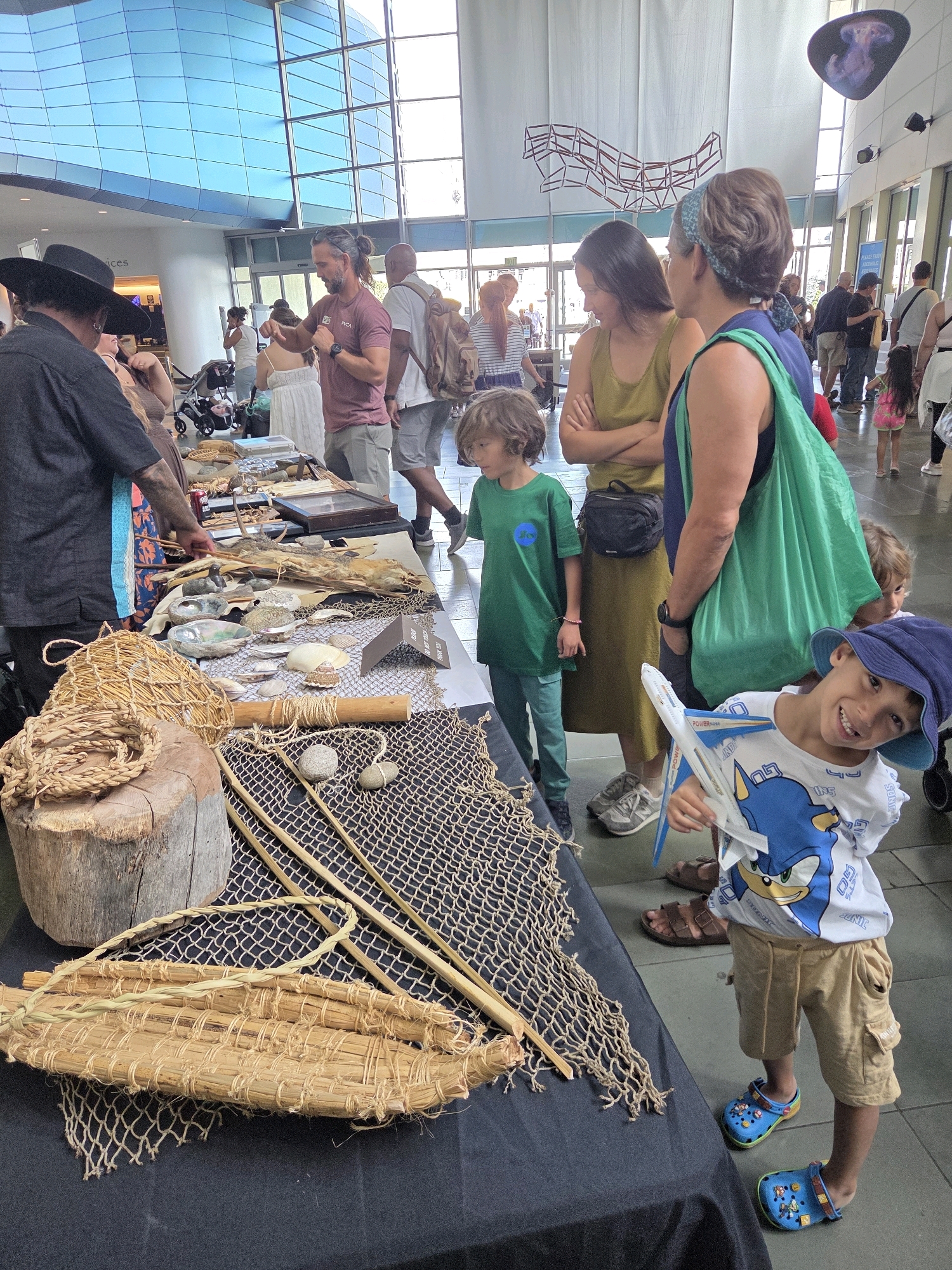 The image depicts an indoor event, likely a marketplace or exhibition, held at the Aquarium of the Pacific in Long Beach, California. The setting features a brightly lit, modern interior with large windows and blue, patterned wall panels. Several individuals are present, browsing or interacting with displays.
In the foreground, a table is laden with various items, including fishing nets, woven baskets, shells, and what appear to be dried specimens or artifacts. A young boy in a blue hat and a t-shirt with a cartoon character is holding a toy airplane. Adjacent to him, another child in a green shirt stands by the table.
Further back, a group of adults are observed. One man in a black hat is looking towards the table. Several women and men are engaged in conversation or examining the merchandise. A stroller is visible near a man with a backpack. The overall atmosphere suggests a casual, family-friendly event.