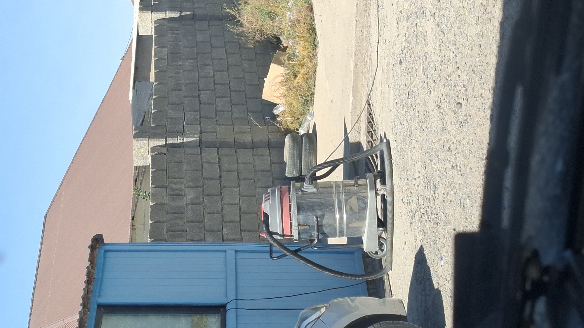 The image depicts an outdoor scene in Шұбарсу, Kazakhstan, likely a service area or yard. In the foreground, a silver car is partially visible on the left. The central subject is a large, metallic, wheeled wet-dry vacuum cleaner with a red and grey top, positioned on a gravel surface. Two car tires are stacked behind the vacuum. Further back, a low wall constructed from gray cinder blocks forms a boundary. Beyond the wall, a building with a reddish-brown, corrugated metal roof is visible under a clear blue sky. To the right of the vacuum, some dry vegetation and a cardboard box are present. The overall environment appears somewhat unkempt, with scattered debris.