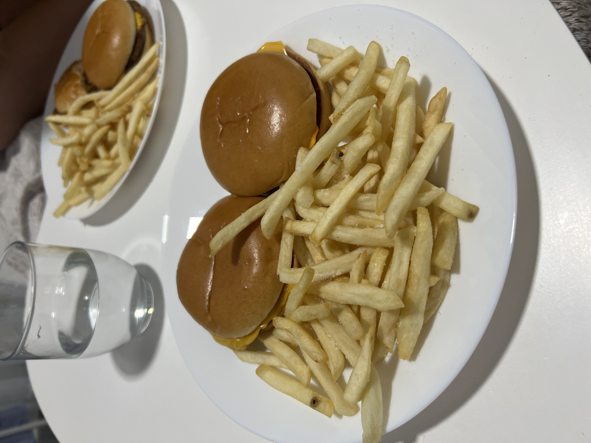 The image shows two white plates of food. The plate in the foreground contains two cheeseburgers and a large pile of French fries. The plate in the background also contains burgers and French fries. To the left of the plates is a partially filled glass of water. The surface beneath the plates and glass is white. The location is unspecified but is unlikely to be in Mogán, Spain.