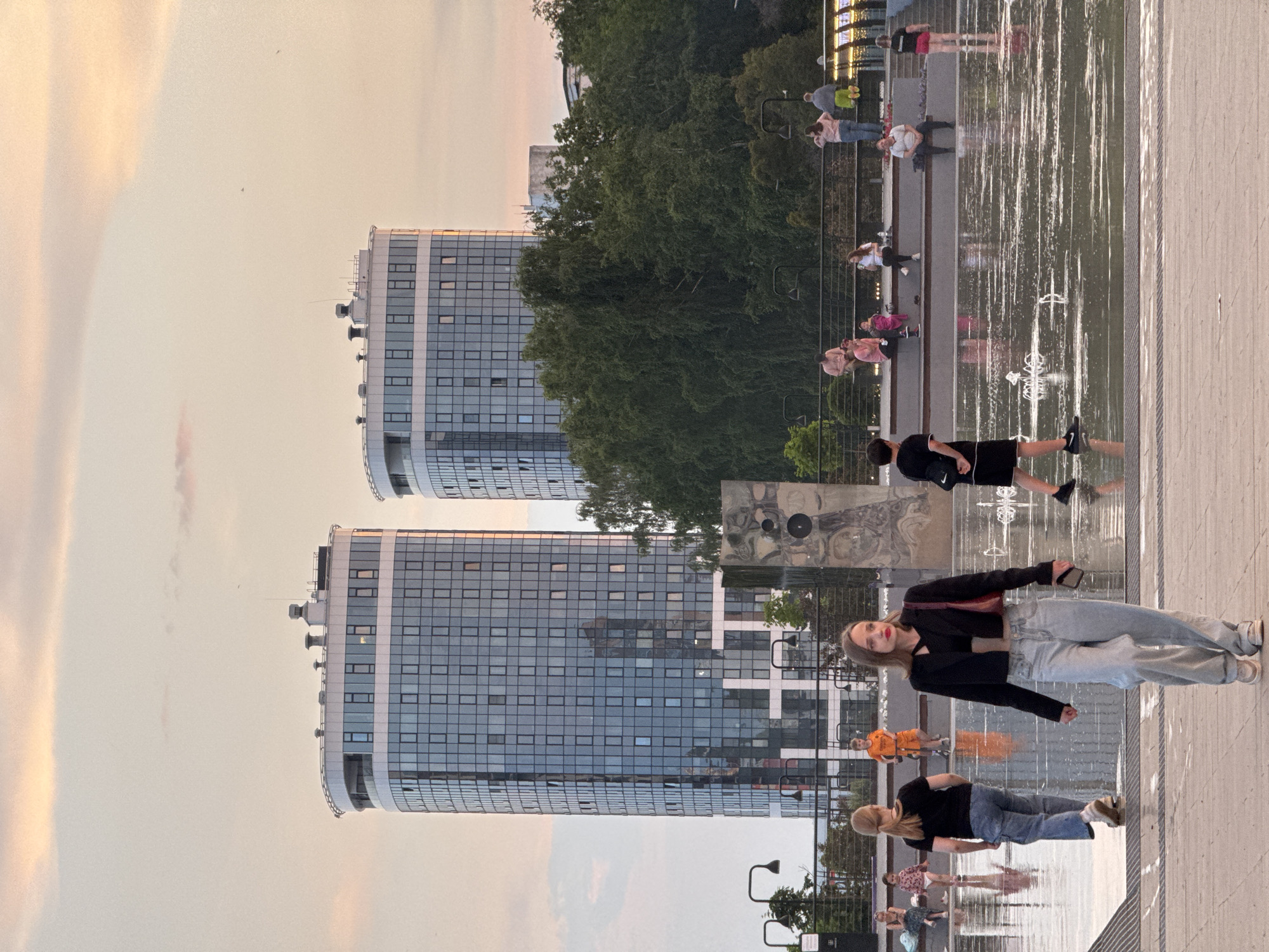 In Izhevsk, Russia, on a cloudy evening with pastel hues in the sky, a group of people are gathered in a modern urban plaza. Two tall, cylindrical glass-fronted buildings dominate the background, their reflections mirroring the sky and surrounding trees. In the mid-ground, a line of lush green trees separates the plaza from the buildings, and a metal structure resembling a sculpture or sound system is visible. The foreground is a wet, reflective plaza, likely a splash pad or fountain area, with several individuals interacting with the water features. A young woman in a black jacket and light-wash jeans walks towards the camera, holding a phone. To her left, a woman with blonde hair in a black t-shirt and jeans walks away from the camera, her back to the viewer. Further back, a young boy in an orange t-shirt and shorts stands near a fountain. Other individuals are seated on tiered steps in the mid-ground, some interacting and others observing the scene. The overall atmosphere suggests a relaxed, community gathering space within a contemporary urban environment.