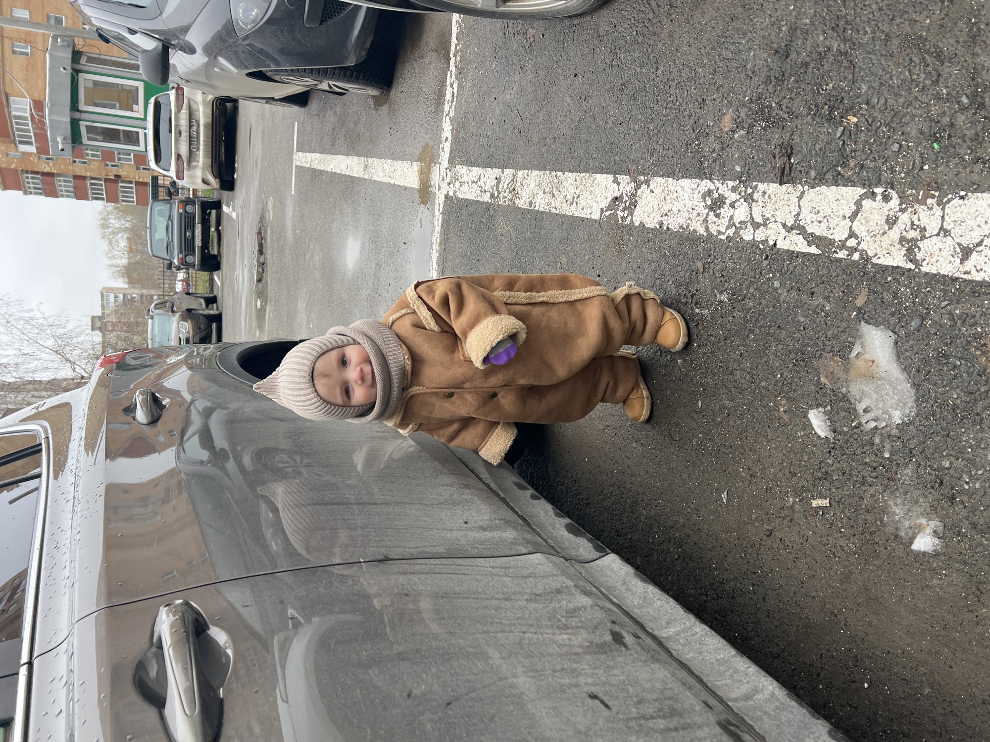 A baby, dressed in a warm, brown, sheepskin-lined jumpsuit and a light beige knit hat, stands on the right side of a wet, dark grey car that occupies the lower left and central portion of the frame. The baby looks towards the camera with a soft smile and holds a small purple object in one hand.
The scene is an outdoor parking lot or street with dark asphalt pavement, marked with weathered white parking lines running horizontally and vertically across the middle of the frame. The ground is wet and contains patches of melting snow or slush, suggesting cold and damp weather conditions.
In the background, several other cars are parked, including a white sedan and a dark-colored SUV (resembling a Lada Niva). Behind the cars, multi-story residential or urban buildings with numerous windows are visible under an overcast sky.
The overall impression is of a casual outdoor moment in an urban environment during a cold, non-sunny day, possibly in late winter or early spring.
The license plate on the white car in the background displays "В135ОХ 797".