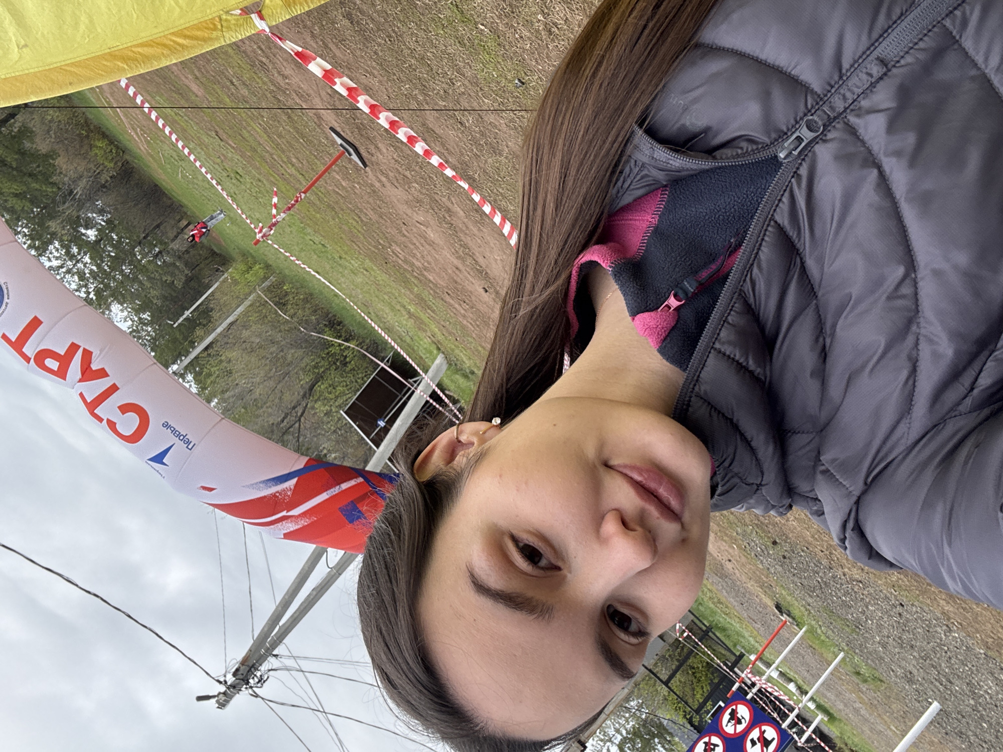 The image shows a selfie of a woman. She has long, brown hair and is wearing a gray puffer jacket with a pink fleece lining. In the background, there is a "START" banner in Russian (translated, it says "First"). There are red and white striped barricade tapes and a grassy field. There are poles with circular signs bearing illustrations of animals. The location is identified by the Cyrillic text on the start banner as Orenburg, Russia. The sky is overcast.