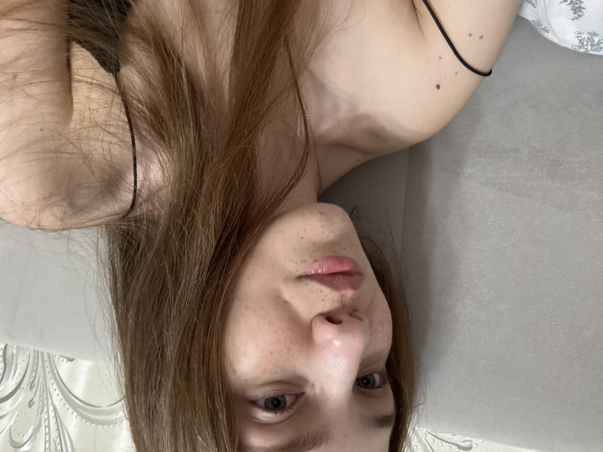 The image shows a close-up, selfie-style view of a young woman with light skin, freckles, and auburn hair. She is lying down, possibly in bed, with a textured gray headboard behind her. She is wearing a thin black strap, possibly from a camisole or bra. The lighting suggests an indoor setting, and the focus is on her face and upper body. There are no discernible landmarks or features to suggest a specific location within Naberezhnye Chelny, Russia.