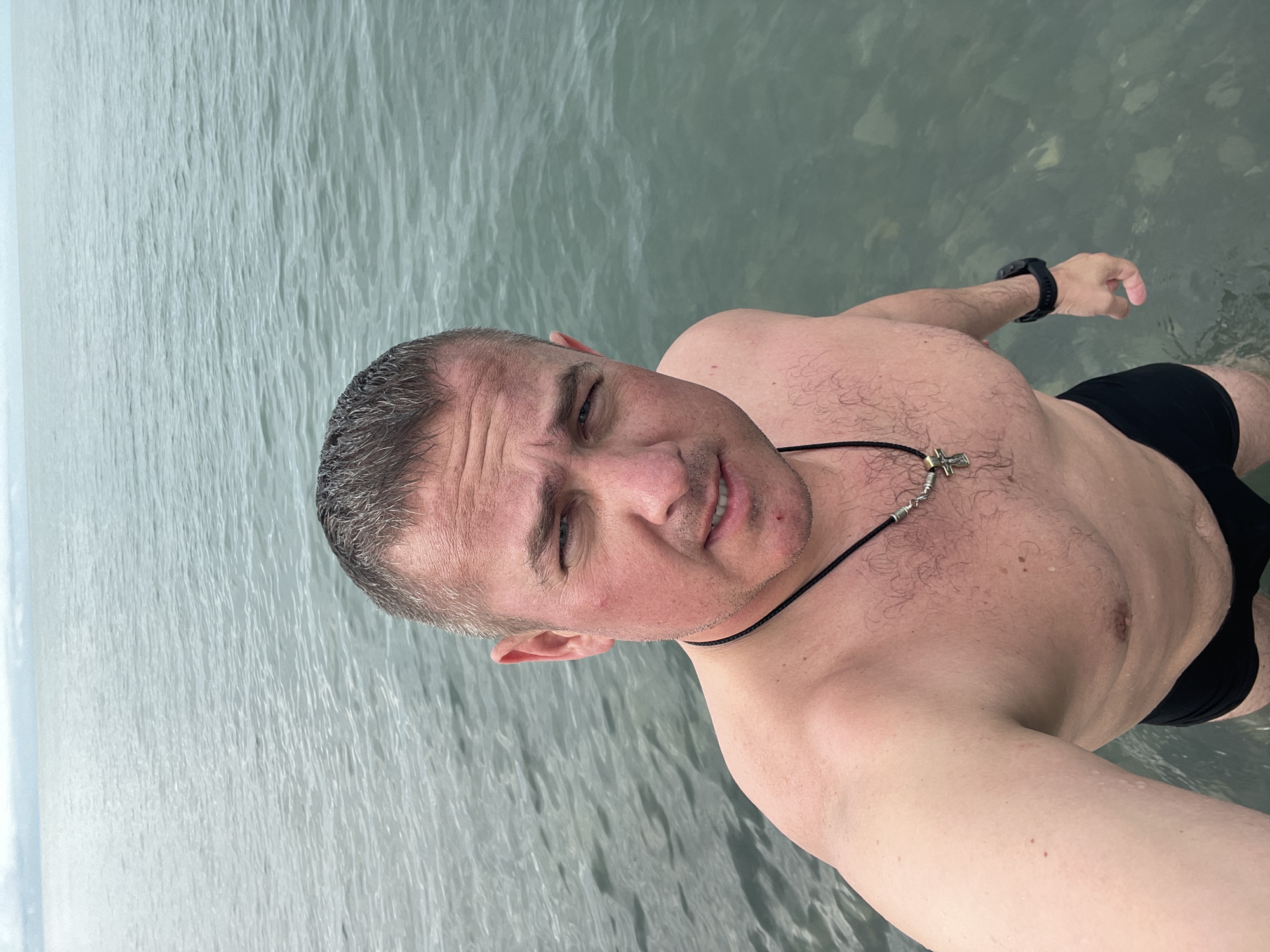 A shirtless man in black swim trunks is standing in water. The man is fair-skinned with short grey hair and is wearing a black watch and a necklace with a cross. The water appears to be a light greyish-green and the background is obscured by the water. The image appears to be taken at eye level by the man himself. It can be concluded from the EXIF data that the picture was taken in Sochi, Russia.
