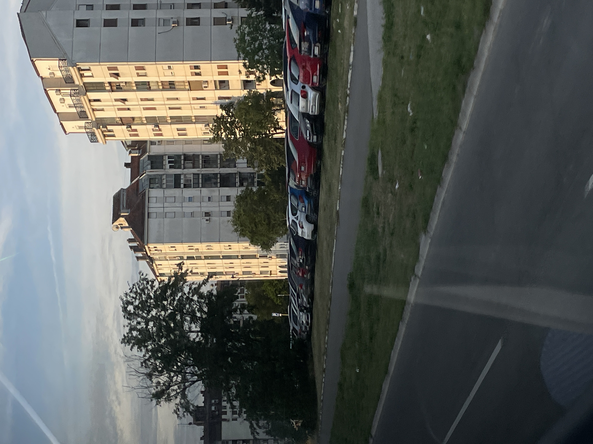 The image depicts a street scene in Belgrade, Serbia. Several high-rise apartment buildings are visible against a pale sky. A line of parked cars is situated along the edge of a paved road, separated by a grassy area. The angle suggests the photo was taken from inside a vehicle.