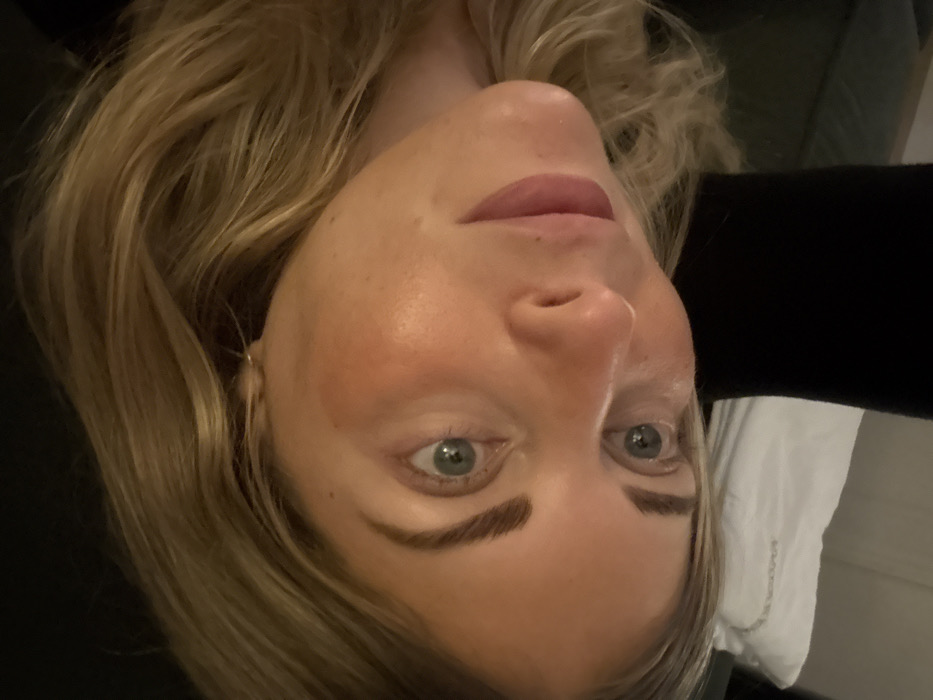 A close-up, high-angle image captures a fair-skinned human subject, appearing to lie down in an indoor setting in Rueil-Malmaison, France. The subject's head is tilted, revealing long, wavy blonde hair framing the right side of the face. Light blue-green eyes, with dark, defined eyebrows, are directed towards the viewer. The nose is central, and the closed lips show a soft pink color, accompanied by a slight blush on the cheeks. The background is blurred, featuring dark fabric to the upper left and a white pillow or folded textile to the upper right. No specific actions or interactions are depicted; the subject is stationary.