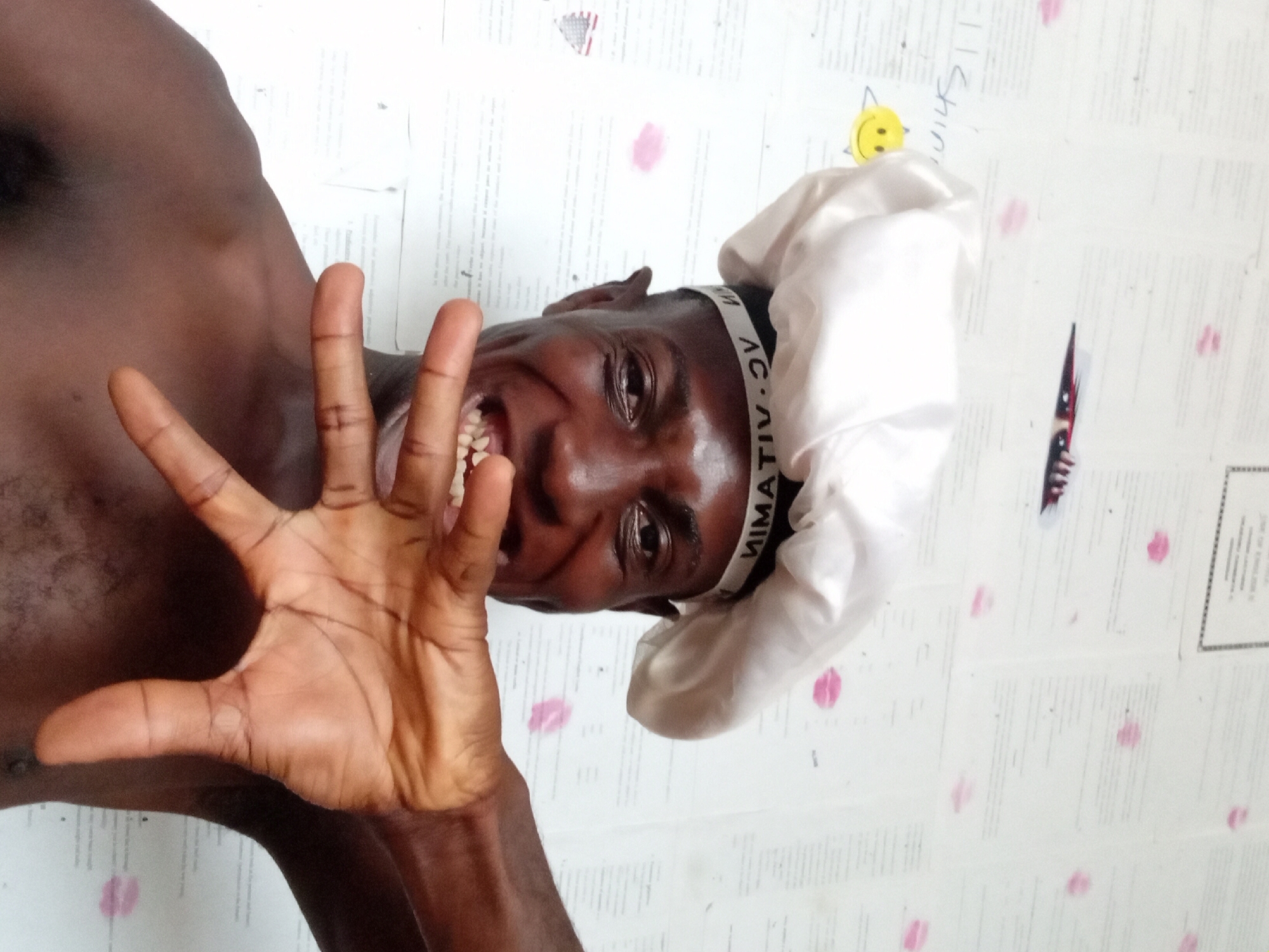The image features a dark-skinned, shirtless person with a white head covering and a black headband, smiling with teeth visible. The person's right hand is extended towards the camera. The background is white with what appear to be printed documents or papers with pink lipstick marks. A sticker of a smiling yellow face is visible in the top right corner. There is no information in the image to confirm the location in the city of Akwete, Nigeria.