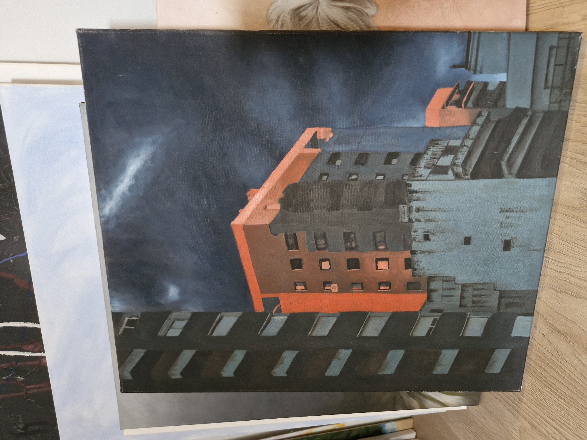 An angled photograph displays a painting, possibly oil or acrylic on canvas, featuring an urban or architectural scene. The painting depicts several buildings rendered in a stylized, somewhat abstract manner, viewed from a high or low vantage point, creating a dramatic perspective.
The dominant colors in the painting are dark blue and grey for the sky, which has a wispy streak of white or light blue, suggesting clouds or atmospheric effects. A prominent building structure is painted in a vibrant, almost neon orange-red, contrasting sharply with the darker tones of the sky and other buildings. Other parts of the architecture are rendered in various shades of grey, dark blue, and light teal, with numerous windows appearing as dark, square, or rectangular openings, and some as elongated slits. The overall impression of the painted scene is one of stark, modern architecture under a dramatic sky.
In the physical photograph, the main painting is placed atop a stack of other artworks or canvases, which are partially visible to the left and beneath. To the right, a wooden surface, likely a floor or table, is visible. In the upper background, out of focus, there is a hint of light-colored hair and a pinkish surface, suggesting a person or fabric in the immediate vicinity. No specific activity is depicted in the photograph or the painting itself. There is no visible text.