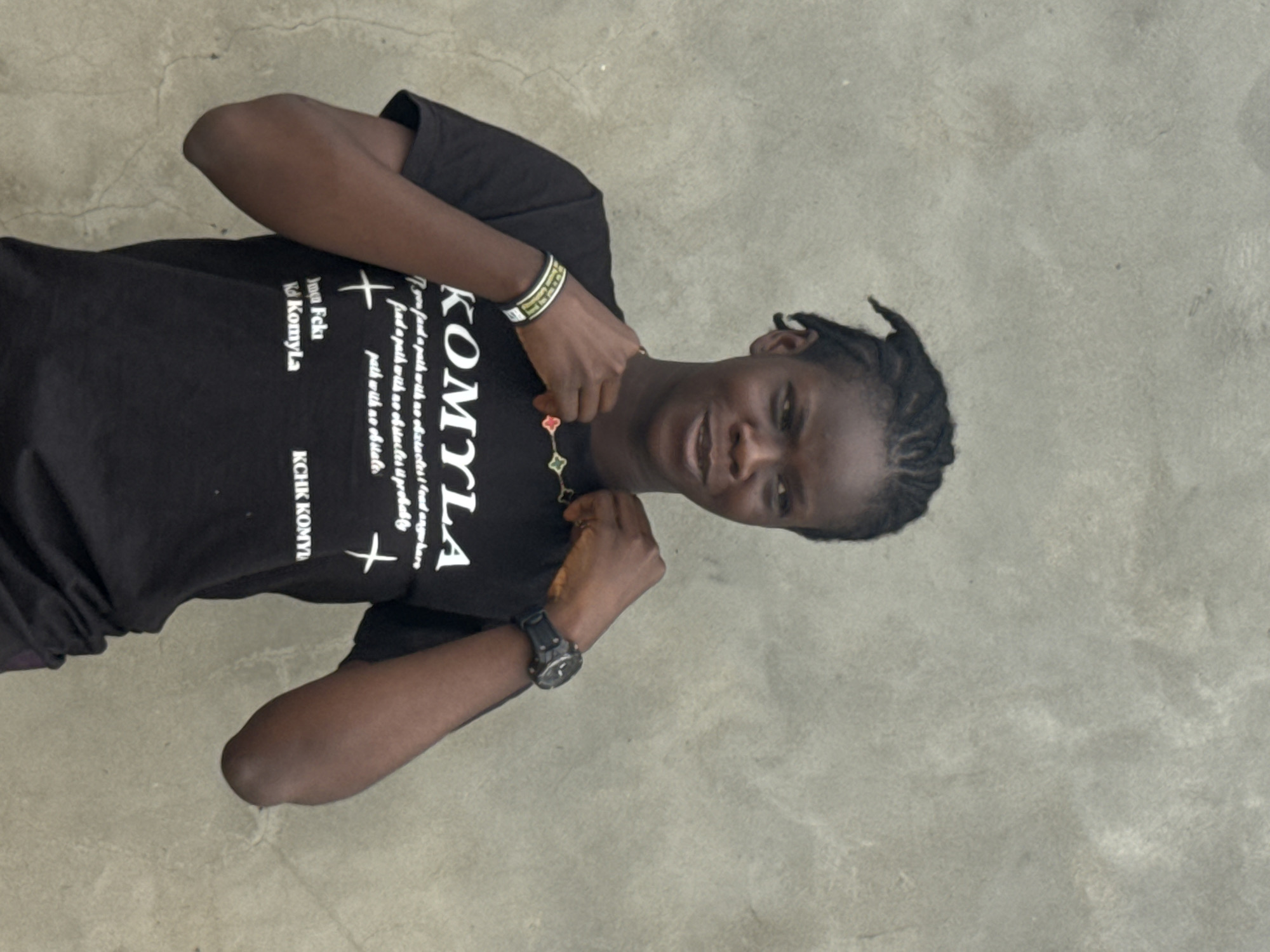 A person is standing on a light gray textured surface. The individual is wearing a black t-shirt with white lettering that includes the word "KOMYLA" and the phrase "you fard a path with no obstacles!" The person has braided hair and is wearing a watch on the left wrist and a bracelet on the right wrist. The person is posing with both hands raised toward their chest. The background consists of the textured surface on which the person is standing. The city of Badagry, Nigeria is not explicitly visible in the image and the location cannot be confirmed based on provided visual evidence.