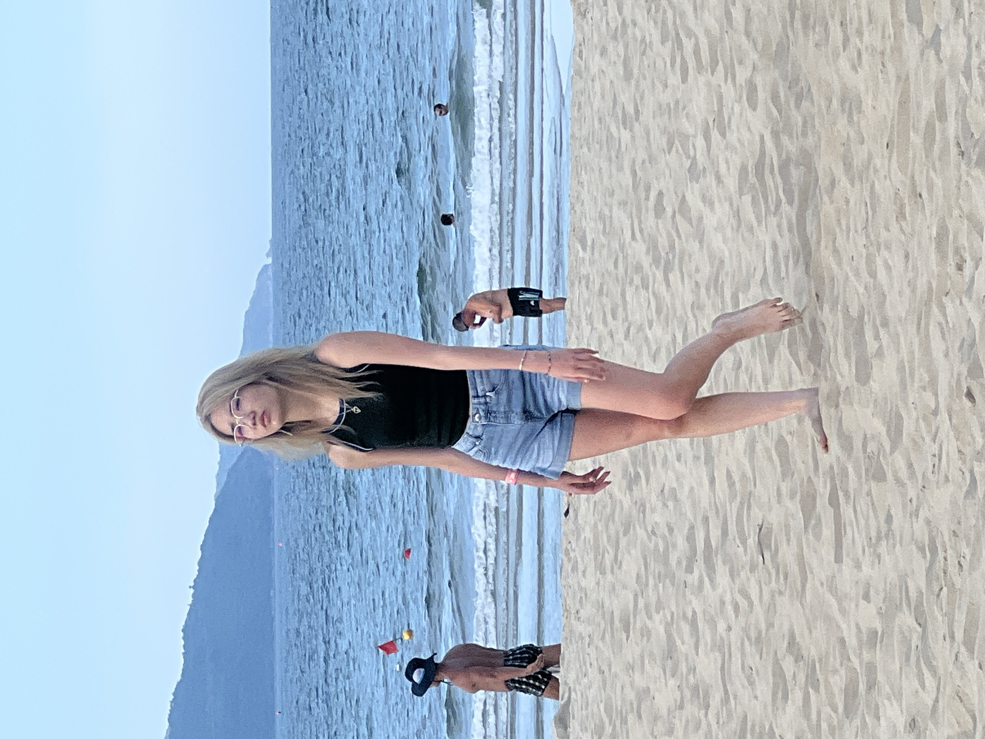 The image depicts a beach scene in Da Nang, Vietnam. In the foreground, a young woman with blonde hair and glasses is walking on the sand, wearing a black tank top and denim shorts. She is looking away from the camera, with her right leg forward. The beach is sandy, with gentle waves lapping at the shore in the background. Several other people are visible in the water. To the left, a man wearing a black hat and checkered shorts stands near the water's edge. Further out in the sea, two men are swimming, with their heads above water. The ocean is a deep blue, and a hazy mountainous landscape can be seen in the distance under a cloudy sky.