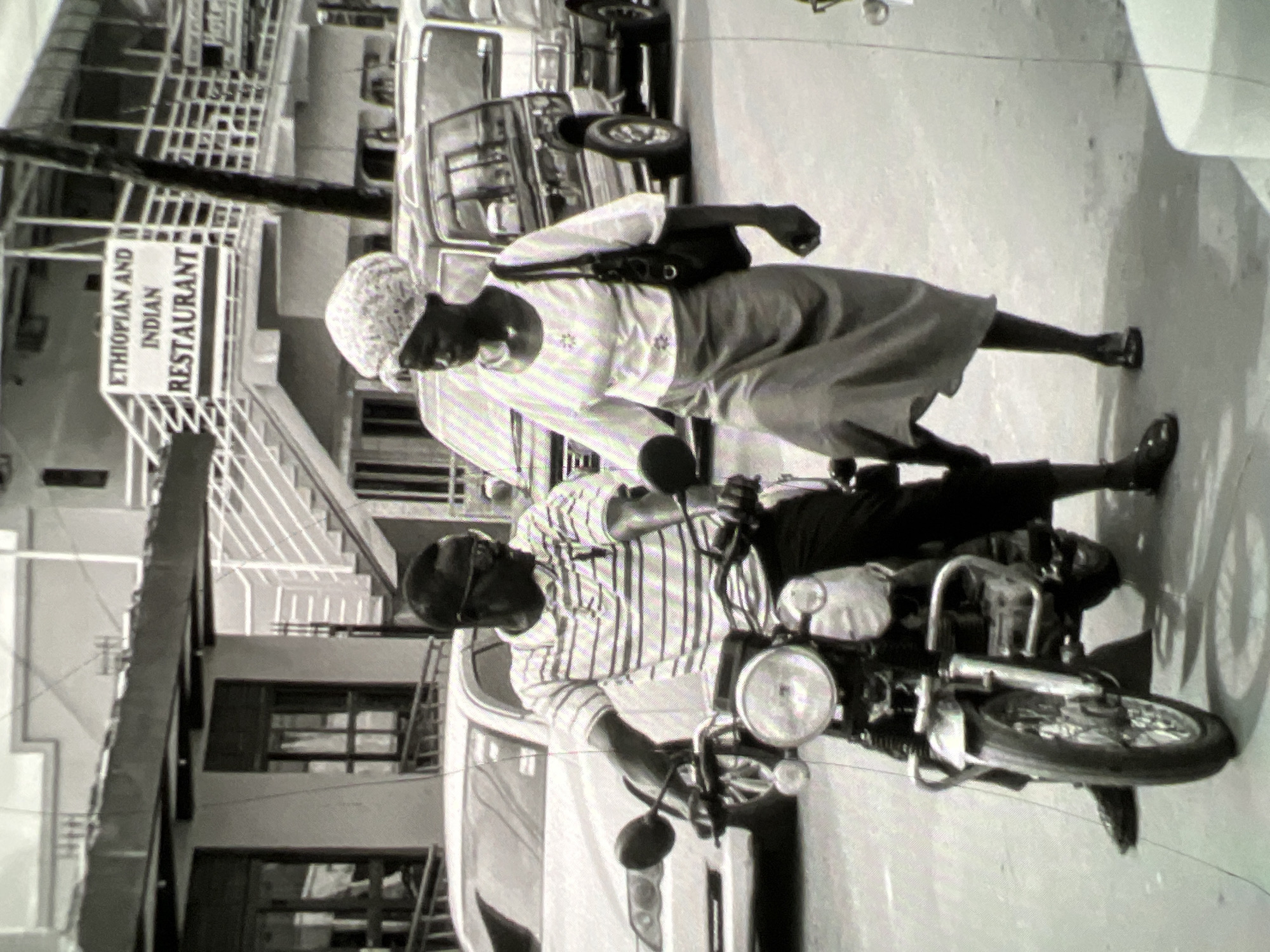 The black and white image depicts a street scene in Lagos, Nigeria. In the foreground, a man wearing glasses and a striped polo shirt is seated on a motorcycle. He is looking to his right. A woman, wearing a headscarf and a long skirt and blouse, is walking next to him and also looking to her right. She has a bag slung over her shoulder. Behind them, there is a white car, and further back, a dark-colored SUV and a white van. In the background, a multi-story building with stairs leading up to it is visible, with a sign that reads "ETHIOPIAN AND INDIAN RESTAURANT." The overall environment appears to be an urban setting with traffic and pedestrians.