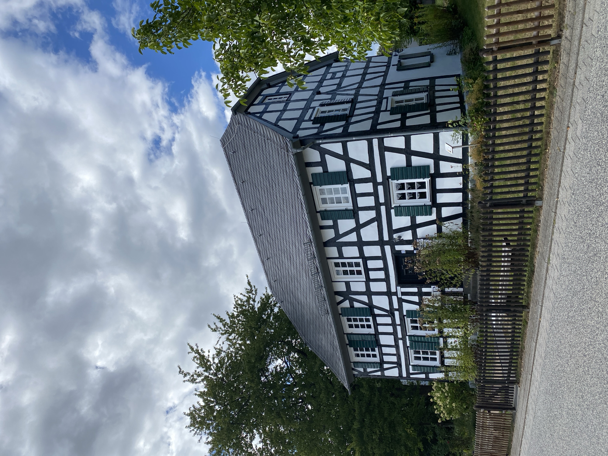 A half-timbered house with a dark grey tiled roof stands on the side of a grey asphalt road. The house's facade is white with black timber framing. It has several windows with green shutters. A dark brown wooden fence runs in front of the house, separating it from the road. Lush green trees and bushes surround the house and fence. The sky above is partly cloudy with patches of blue. The scene is located in Flammersfeld, Germany.