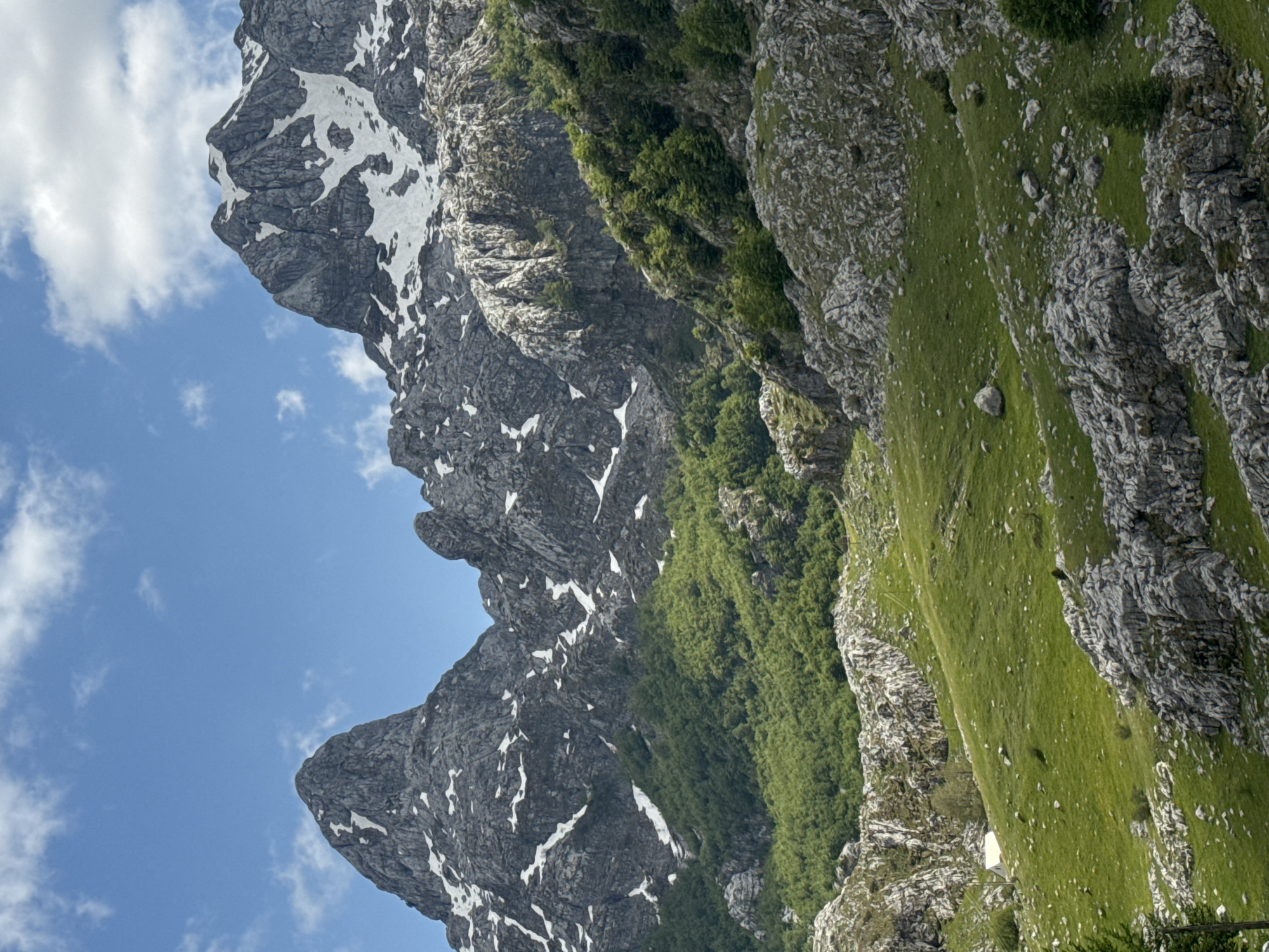 The image shows a mountain range in Brskut, Montenegro. The mountains are rocky, with some green vegetation and patches of snow. The sky is blue with white clouds.
