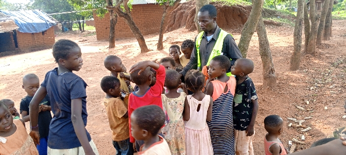 An adult male stands amidst a group of approximately 15-20 children in an outdoor, unpaved area in Ngalula, Democratic Republic of the Congo. The male, with dark skin and short dark hair, wears a dark long-sleeved shirt and a yellow-green reflective vest, possibly displaying "T-C" or "J-C" on the back. He faces the children, looking downwards, with his left hand near the head of a child. The children, of varying young ages, are predominantly dark-skinned and wear diverse casual clothing including dresses, t-shirts, and shorts. They are gathered in a loose semicircle around the adult, with many observing him. The immediate ground is reddish-brown earth, appearing dry. In the background, several single-story structures constructed from red-brown bricks or mud are visible, one featuring a blue tarp on its roof and another with a corrugated metal roof. A line of slender trees with light-colored bark extends across the middle ground to the right. On the far right foreground, discarded corn cobs are scattered on the ground. Green foliage and a small dirt embankment are present in the distant background. The scene is illuminated by natural daylight.