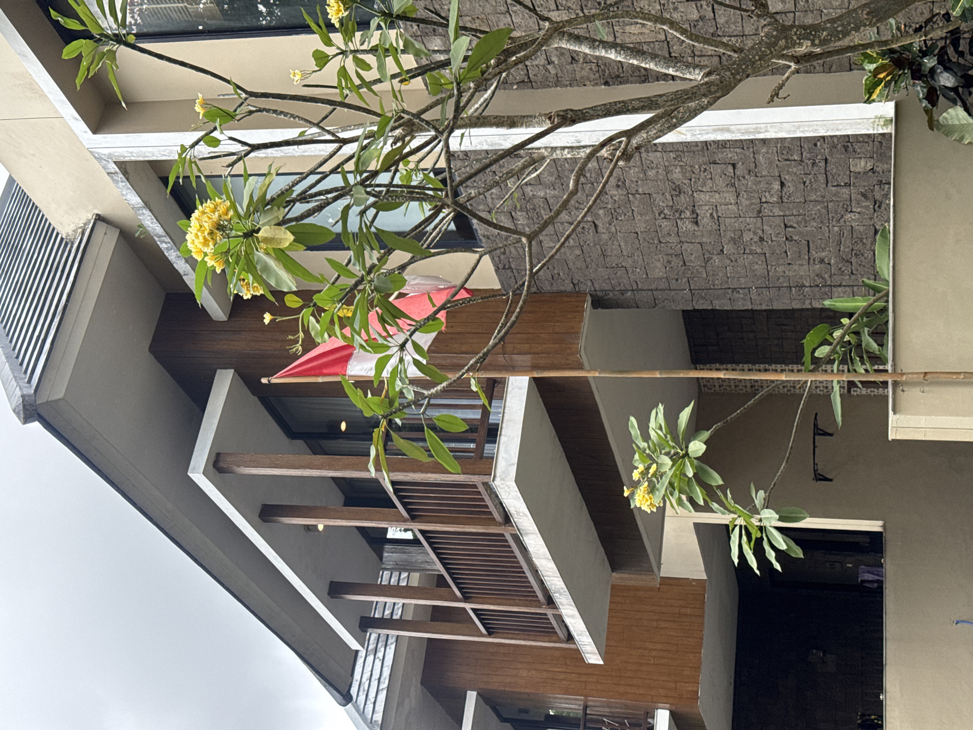 The image shows a close-up view of a building facade with a balcony, a flag, and a flowering tree. The building appears to be a modern structure with a combination of wood paneling, stone accents, and light-colored concrete. A balcony with a wooden railing extends from the second story. A small Indonesian flag, red and white, is planted in front of the building and extends upwards alongside a tree. The tree has a bare, branching structure with clusters of yellow flowers and green leaves. The background is dominated by the building and a cloudy sky. The setting is likely a private residence or a commercial establishment in a tropical or subtropical region, given the presence of the flowering tree and the architecture.