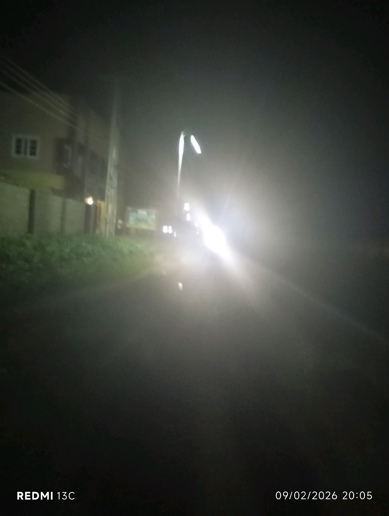 The image depicts a street scene at night, likely in Shendam, Nigeria. A bright light source, possibly a street lamp or vehicle headlights, illuminates the right side of the frame, creating a significant glare and obscuring details in that area. On the left, a building with visible windows is partially visible. A grassy area is present between the building and the street. The road surface is dark and appears wet or reflective. The overall impression is of a dimly lit, hazy urban environment at night. The timestamp in the bottom right corner reads "09/02/2026 20:05". The watermark "REDMI 13C" is in the bottom left corner.
