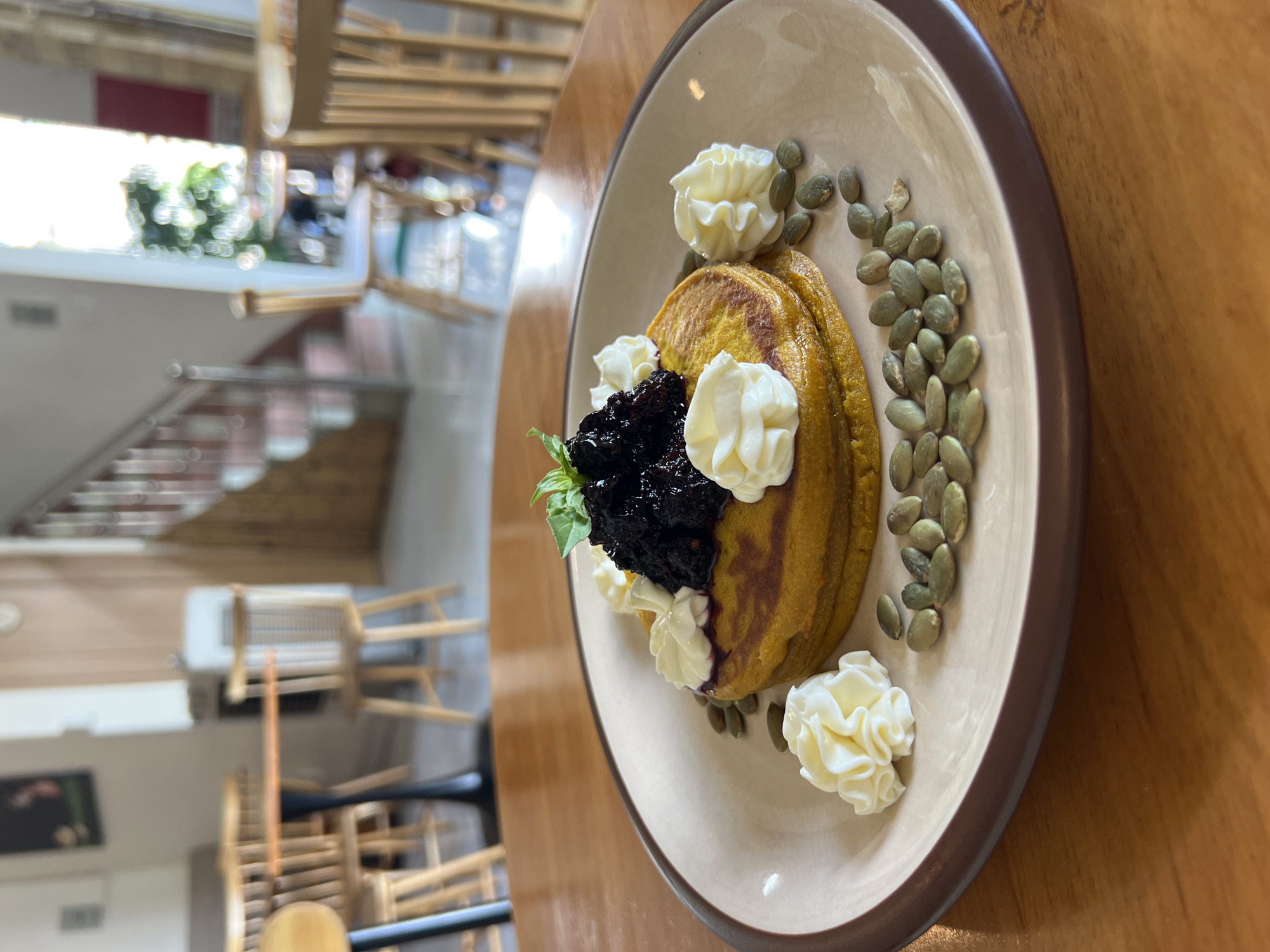 The image shows a plate of food, likely pancakes, topped with dark fruit compote, a mint leaf, and piped cream, alongside a scattering of pepitas (pumpkin seeds). The plate has a light beige base and a dark brown rim, sitting on a wooden table. In the blurry background, there's a view of a building interior, featuring wooden chairs and a staircase. Based on the provided information, the location is in Nha Trang Ward, Vietnam.