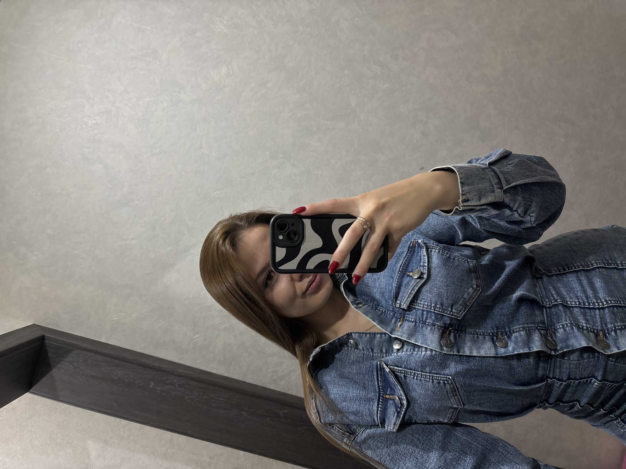 A young woman with long, brown hair is taking a selfie in a room. She is wearing a denim jacket and a denim dress. The room has a textured, light-colored wall and a dark wooden frame is visible on the left side of the image. The woman is holding a smartphone with a black and white patterned case, and her nails are painted red.