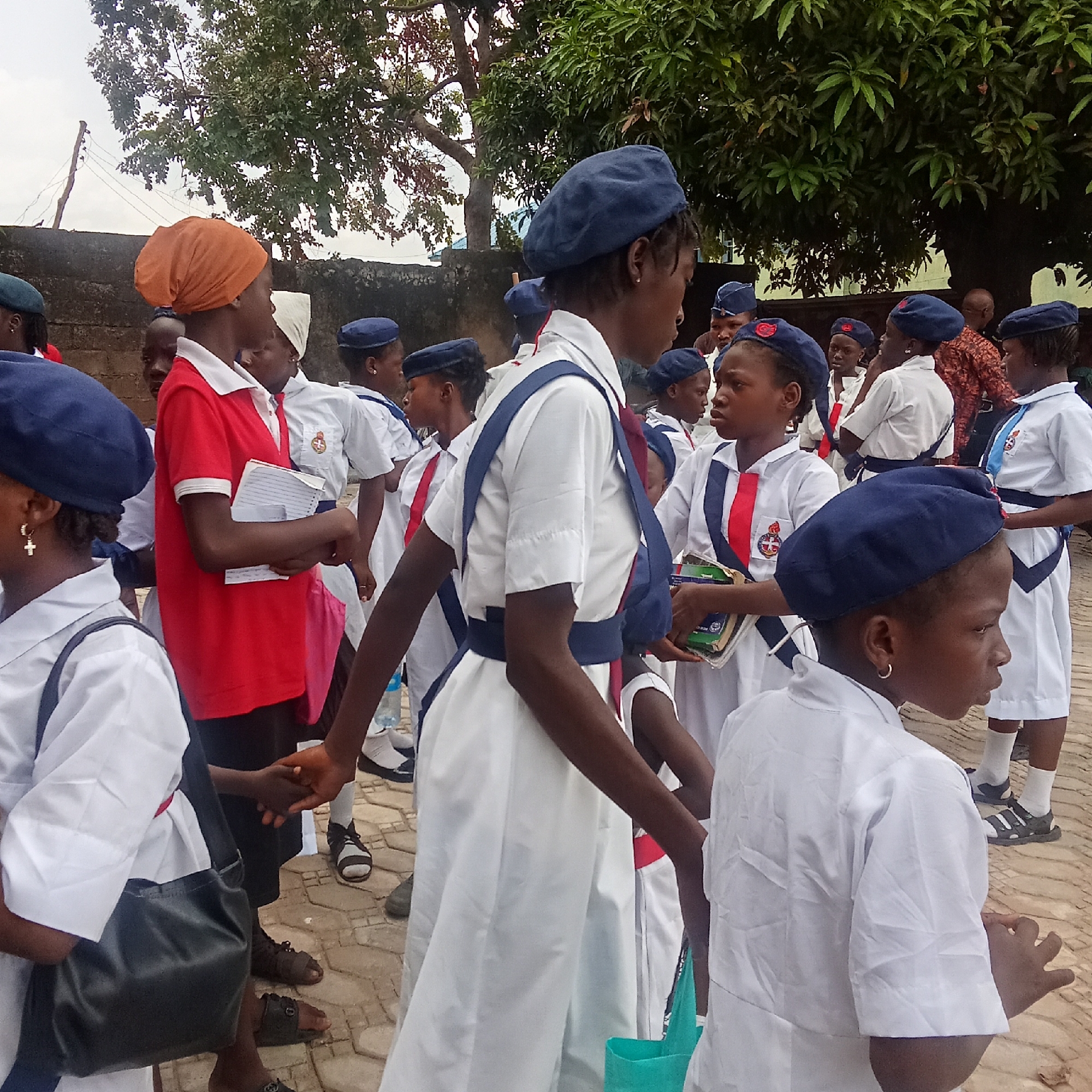 In an outdoor scene likely located in Akwanga, Nigeria, a group of young girls, numbering around fifteen, are gathered. They are predominantly dressed in similar white uniforms accented with red and blue detailing, and navy blue berets. The attire suggests a possible school or organization affiliation. Some of the girls are holding books or notebooks. A few individuals in the background appear to be adults, one wearing a patterned shirt. The setting is likely a courtyard or outdoor space with brick or paved ground. Trees provide a backdrop, contributing to an open-air environment. The overall scene has the air of a gathering or meeting.
