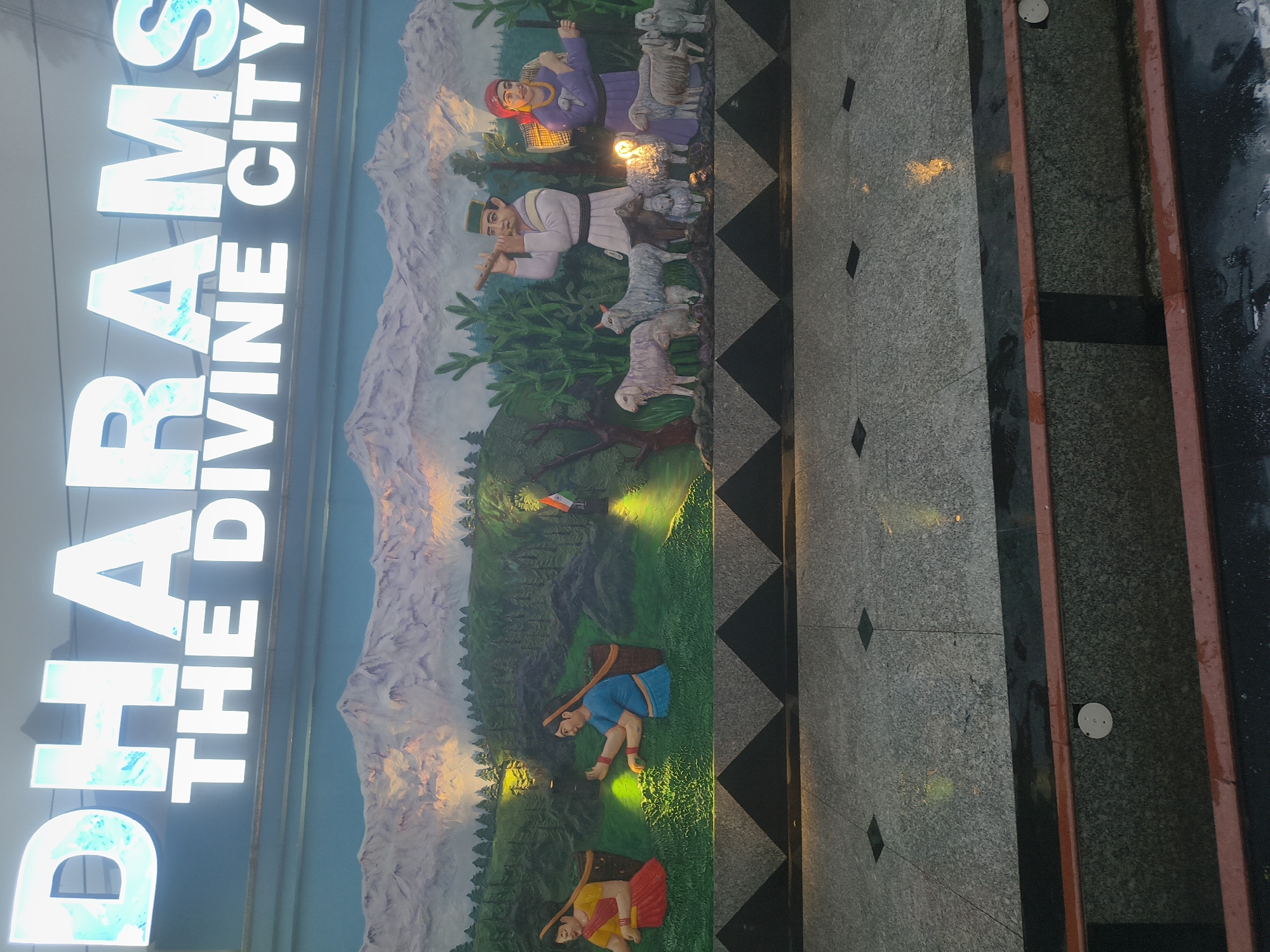 The image shows a large, illuminated sign that reads "DHARAMS THE DIVINE CITY" in prominent, white and light blue letters. Below the sign, there is a mural depicting a scenic mountain landscape with a tea plantation on the left, featuring two women in traditional attire harvesting tea. To the right of the tea plantation, the mural continues with a scene of a man playing a flute and a woman carrying a basket, accompanied by several sheep. A small Indian flag is visible in the tea plantation section. The mural is set against a backdrop of snow-capped mountains. The foreground shows a stone-tiled walkway with decorative triangular patterns and subtle lighting. The overall impression is of an entryway or prominent signage for a city or tourist destination.