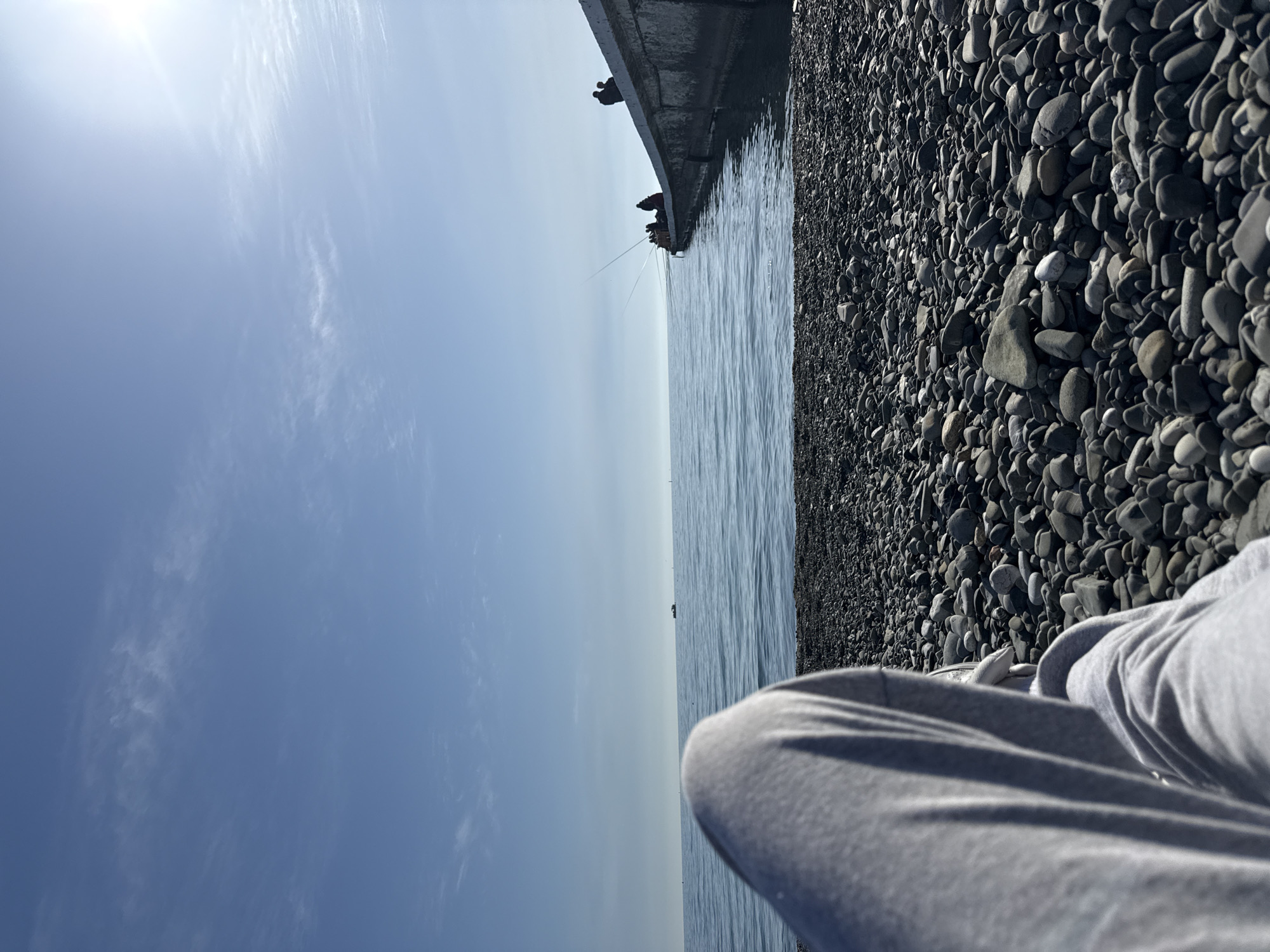 A first-person perspective image captures a sunny day at a rocky beach. In the foreground, the lower legs of a person, clad in grey sweatpants, rest on a dense expanse of dark pebbles and stones that cover the right side of the frame. To the left, the calm, blue-grey water of a sea or large lake stretches horizontally. A concrete pier or wall extends into the water from the right, where several silhouetted figures, likely fishermen, are visible sitting or standing. Multiple fishing rods extend from the pier into the water. The upper half of the image features a clear, light blue sky with faint, wispy white clouds. A bright sun glare is prominent in the upper left, indicating a bright mid-day or afternoon setting. The scene suggests a peaceful outdoor activity, possibly fishing or relaxation by the coastline. No visible text is present.