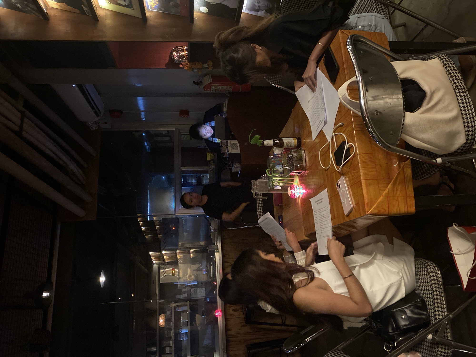 The image depicts a dimly lit indoor setting, likely a restaurant or bar, with a group of people seated around a table. In the foreground, two women are visible, engrossed in documents spread on a wooden table covered with an orange tablecloth. One woman, wearing a white sleeveless dress, holds a document in her hands. The other, wearing a black top, also has papers in front of her. On the table, there are also a bottle of beer, a jug of water with ice, a small plant in a glass, and decorative candle holders.
In the background, another woman in a black shirt stands behind a counter, seemingly attending to a laptop. Further back, a kitchen area is visible with shelves stocked with items. The overall ambiance suggests a casual dining or meeting environment. Based on the context provided, this scene is situated in Davao City, Philippines.