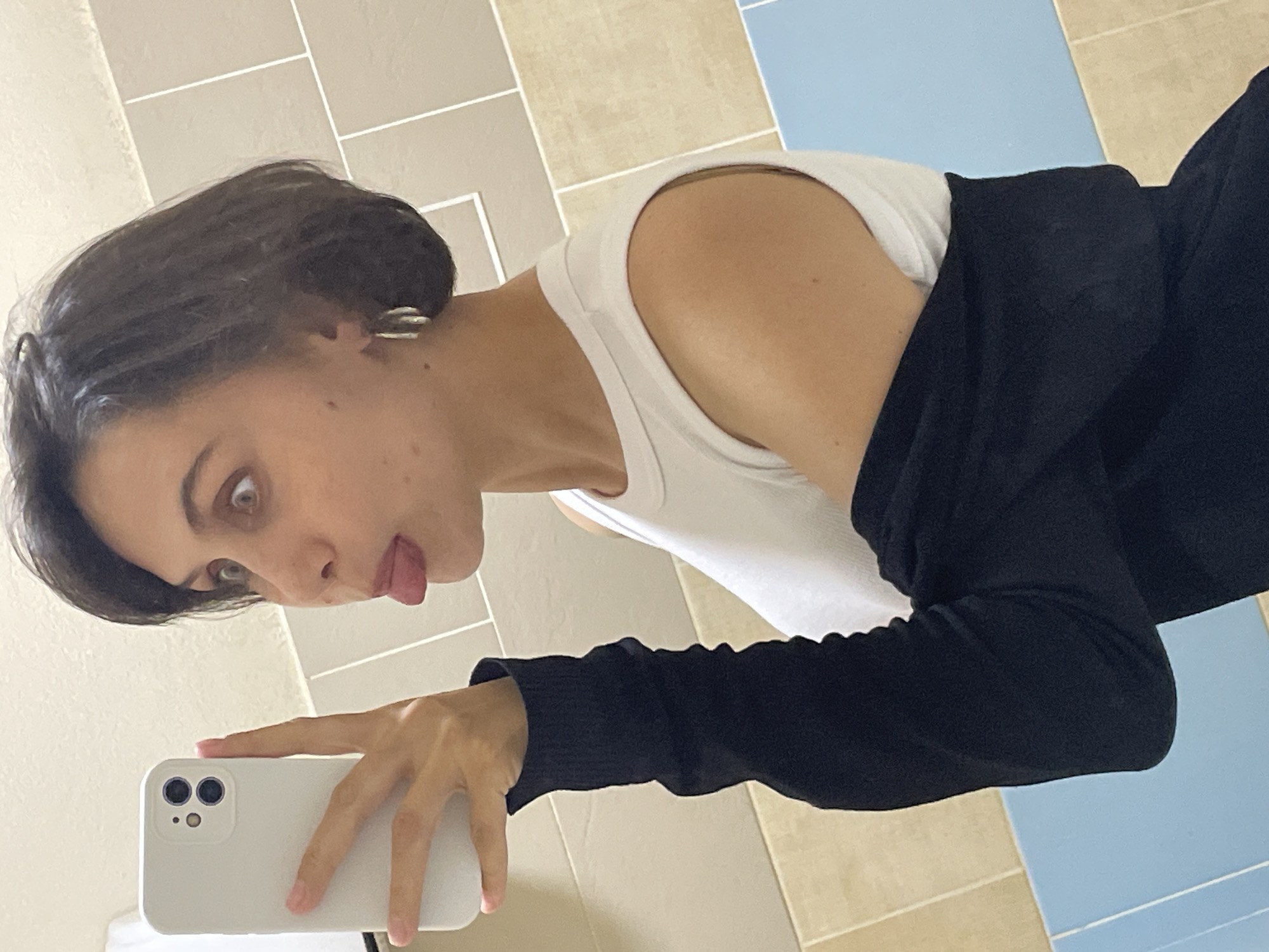 A young woman is taking a selfie in a bathroom. She is holding a white smartphone, presumably taking the photo. She is sticking her tongue out and looking into the camera with a playful expression. She is wearing a white tank top with a black sweater or cardigan over it. Her hair is short and dark. The background consists of beige and blue tiles, suggesting a bathroom setting. There is no information available that indicates this image was taken in Izhevsk, Russia.