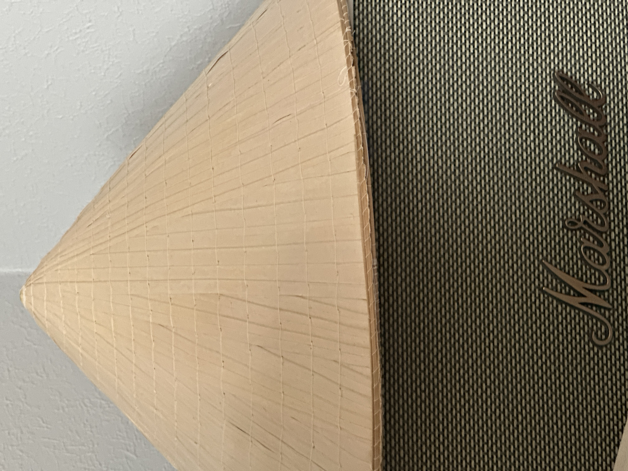 The image shows a close-up view of a conical hat, likely a traditional Asian conical hat made of woven straw or bamboo, resting on top of a Marshall amplifier. The hat is light brown in color and displays a visible weave pattern with horizontal and vertical stitching. The amplifier below is black with a textured grille and features the prominent "Marshall" logo in a metallic cursive font. The background is a plain white, textured wall. There are no people or animals visible in the image. The scene appears to be an indoor setting, possibly a room or studio. The image is cropped to focus on the hat and the amplifier, with the wall providing a simple backdrop. There is no information within the image or its context to suggest a location in Nevinnomyssk, Russia.