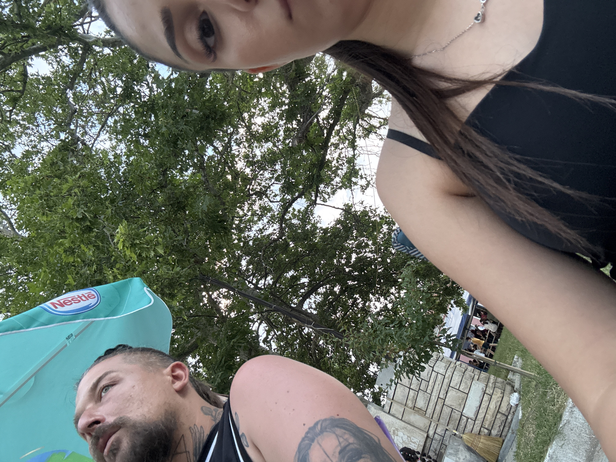 A selfie-style image captures two individuals in an outdoor setting in Kostievo, Bulgaria, beneath a canopy of green trees and a partial view of a blue sky. On the left, a male with a beard, mustache, and visible tattoos on his arm and neck/shoulder looks upwards to his left. He wears a black tank top. A light blue umbrella, with a "Nestlé" logo visible on its underside, partially shades him. On the right, a female, presumably taking the photograph, shows the right side of her face, right shoulder, and upper torso. She has dark hair tied back and wears a black strap top with a silver necklace featuring heart-shaped pendants. In the background, partially obscured by foliage and a stone-clad wall with a broom leaning against it, several indistinct figures are gathered, suggesting a social event or market. The ground shows grass and dirt.