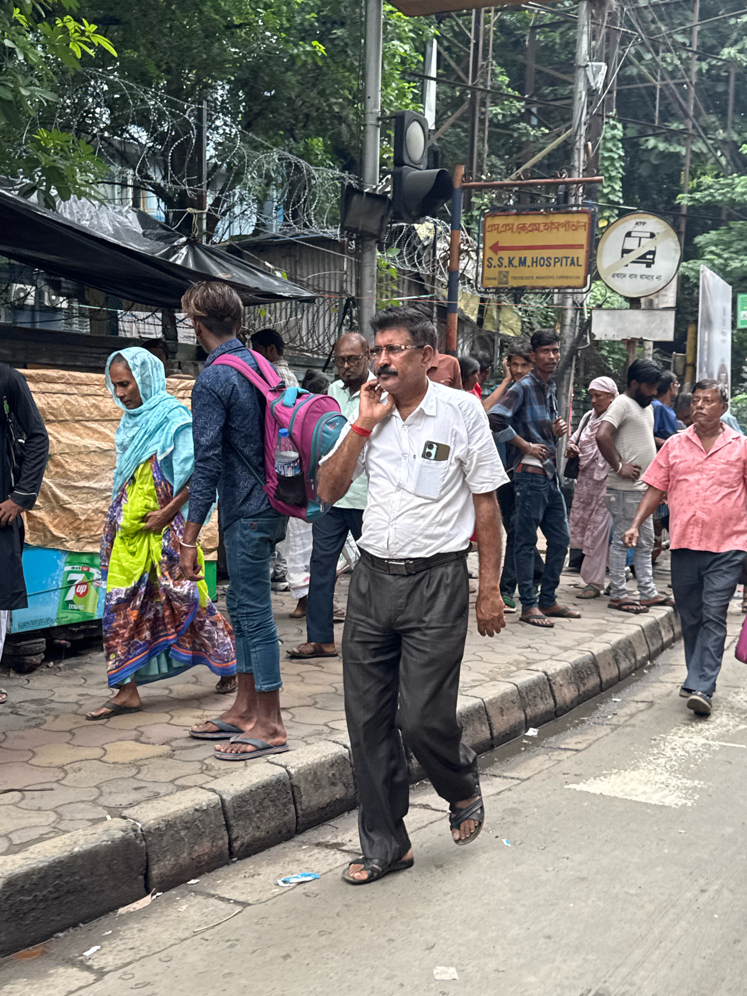 The image depicts a street scene in Kolkata, India, near SSKM Hospital. A man in a white shirt and dark pants is walking and talking on his phone. A woman in a blue headscarf and a colorful sari walks past him, carrying a bag. Behind them, a young man with a pink backpack walks on the sidewalk, which is lined with a curb. In the background, a traffic light is visible, along with various signs, including one indicating "S.S.K.M. HOSPITAL". Barbed wire is strung along a fence on the left side of the image. The overall environment suggests a busy urban area with pedestrian traffic and an overcast sky.