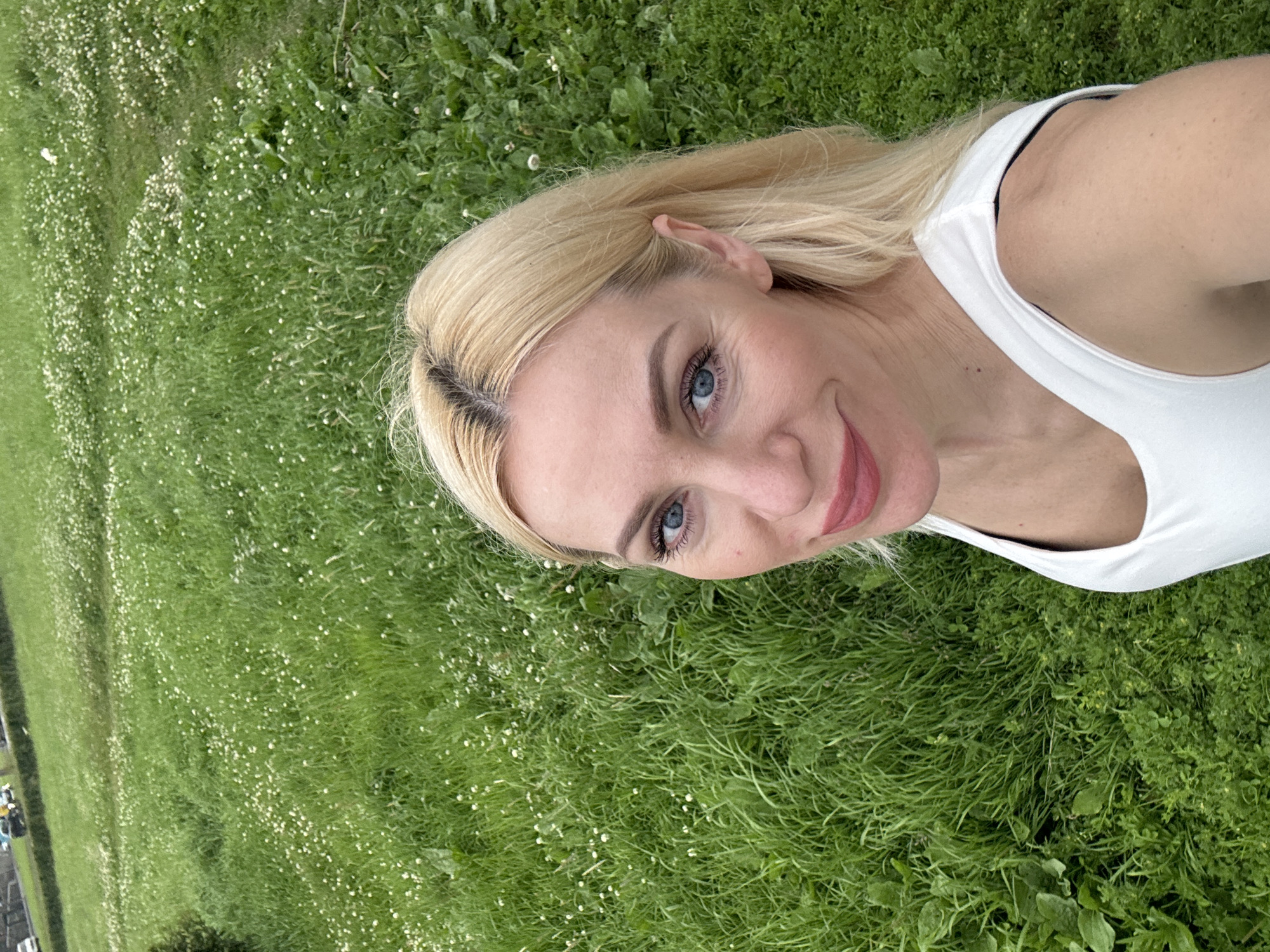 A blonde woman in a white top smiles for a selfie in front of a grassy field. The field is covered in green grass and small white flowers. In the background, there is a road with a few cars on it. The woman has blue eyes and pink lipstick.