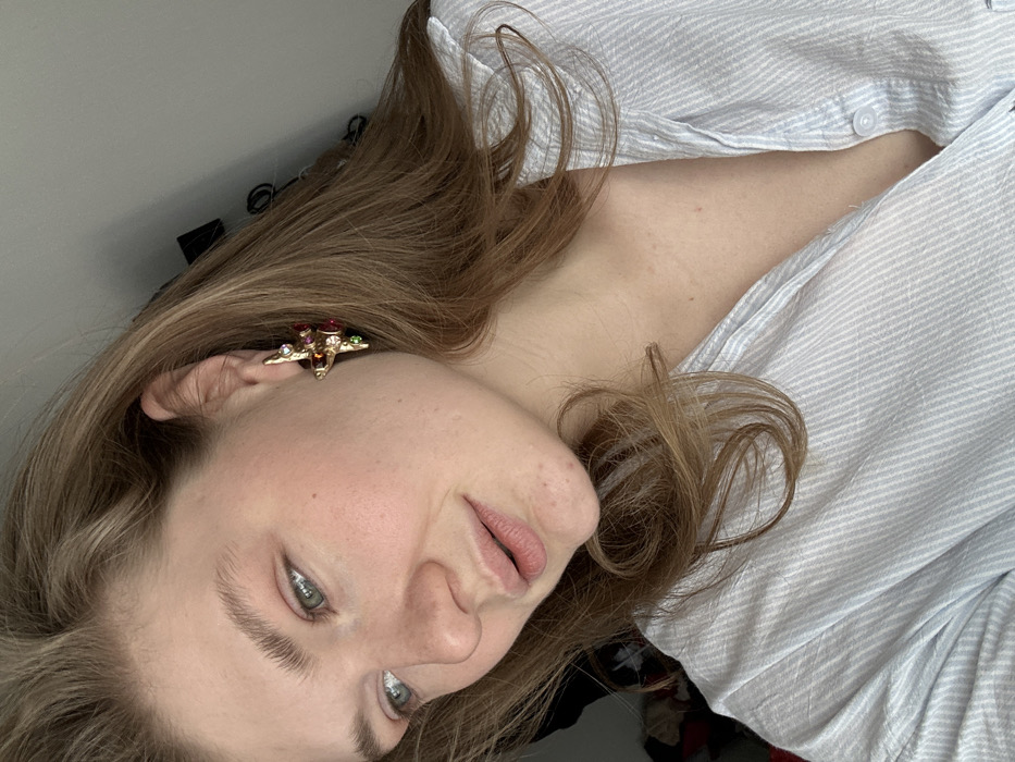 A medium close-up photograph captures a young adult female indoors, posing for the camera. She has fair skin, light brown hair parted on the left side, and light-colored eyebrows. Her eyes, with visible green-blue irises, are directed towards the left side of the frame. Her lips are light pink and slightly parted. A golden, multi-gemmed earring, possibly depicting a stylized airplane or cross with red, green, and clear stones, is visible on her right earlobe. She is wearing a light blue and white pinstriped button-up shirt, partially unbuttoned, revealing a portion of her upper chest. The background is a uniformly lit, indistinct interior space, with a dark, possibly black, object faintly visible in the upper right corner. No identifiable geographical markers for Kazan, Russia, are present within the visual information provided by the image.