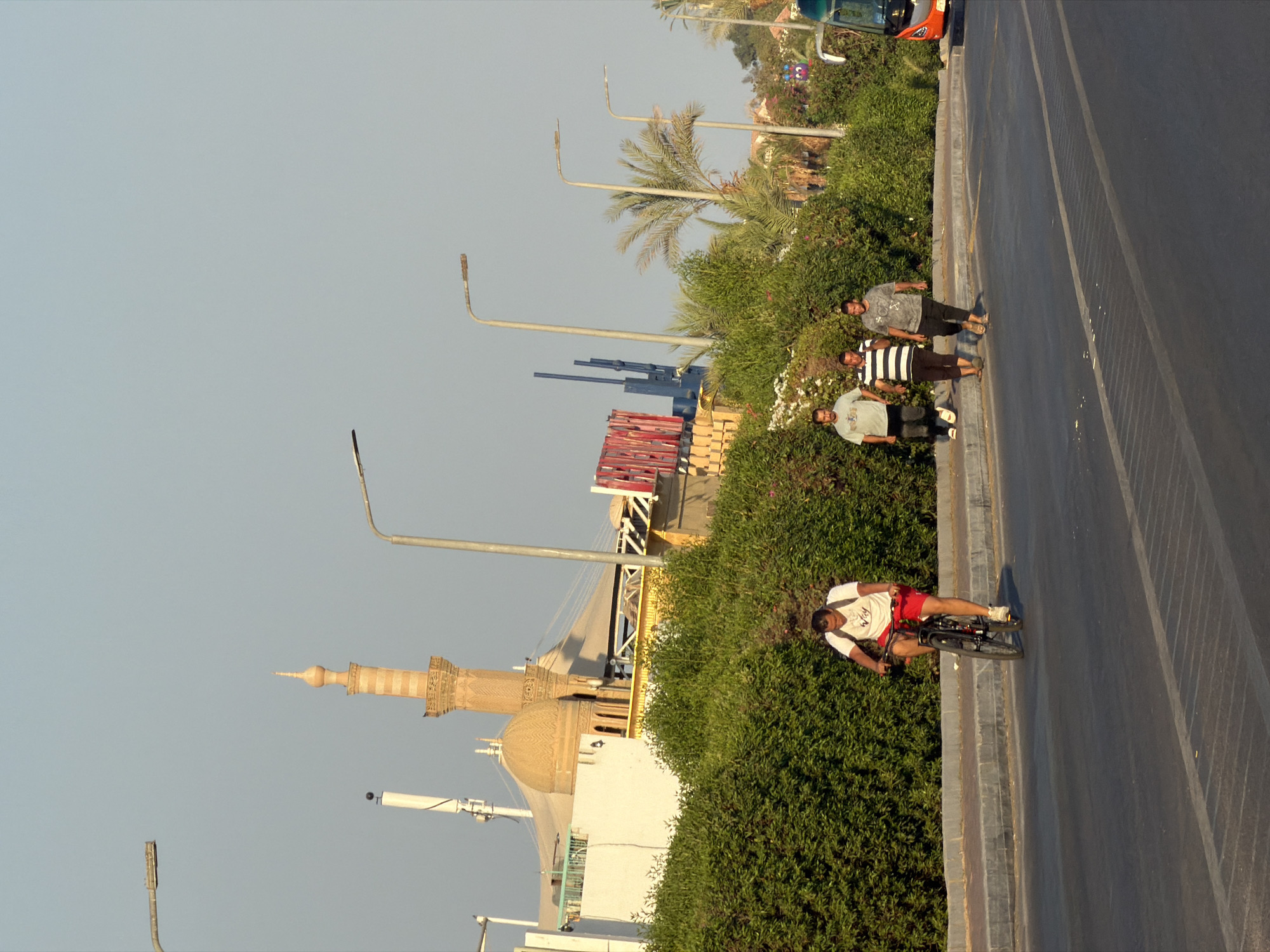 The image depicts a street scene in Sharm El Sheikh, Egypt. In the foreground, a man is riding a bicycle on a paved road. He is wearing a white t-shirt and red shorts, and is carrying a backpack. To the right of the cyclist, a group of three men are walking on a sidewalk. They are dressed in casual clothing. Behind the sidewalk, there is a dense hedge of green bushes. In the background, a minaret of a mosque with a dome and a spire rises above the buildings. Further in the background, palm trees and other vegetation are visible, along with a bus on the right side of the image. The sky is clear and pale blue. The overall scene suggests a warm, sunny day in an urban environment.